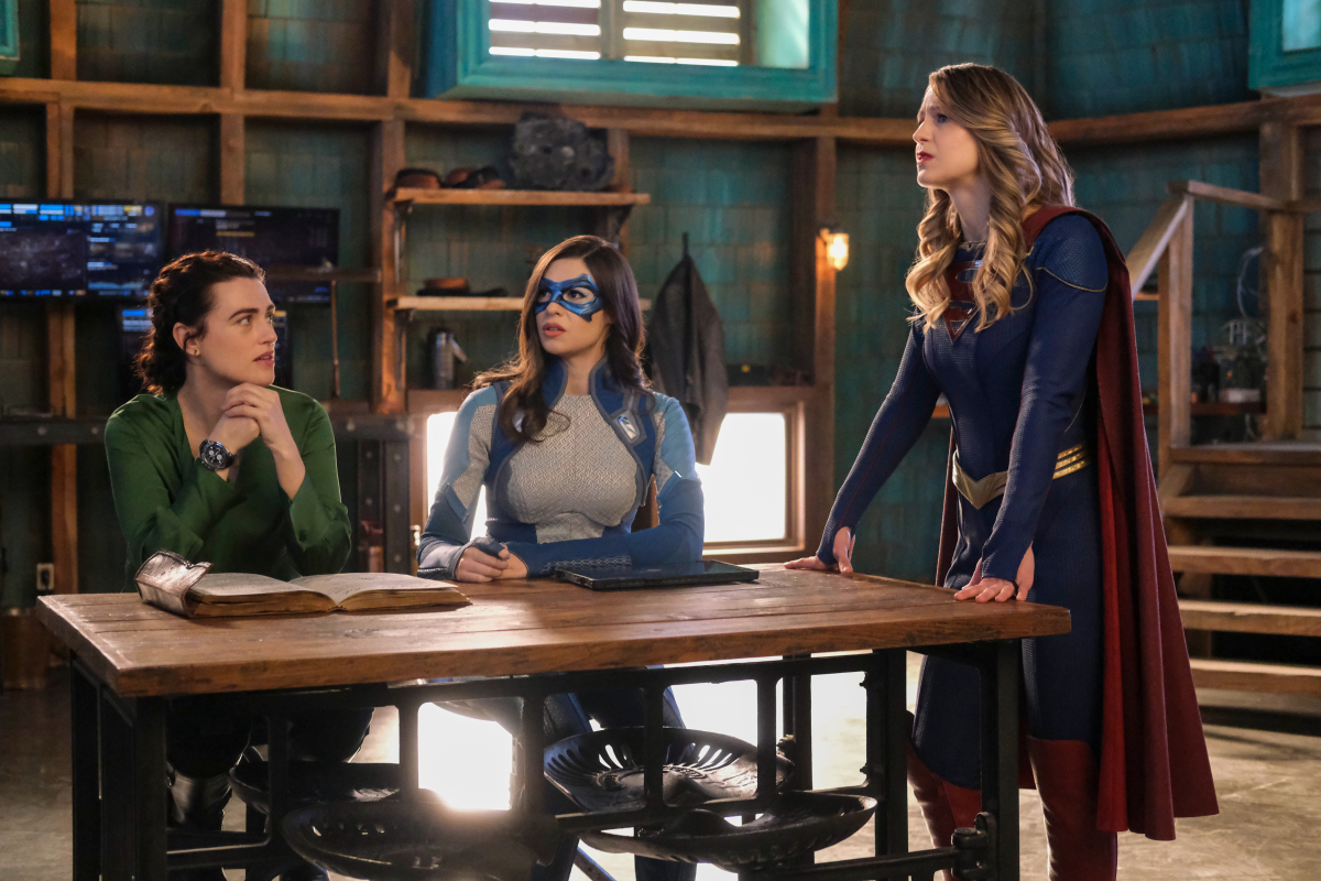 'Supergirl' Season 6 Episode 14 actors Katie McGrath, Nicole Maines, and Melissa Benoist, in character as Lena Luthor, Dreamer, and Supergirl, gather around a table. Dreamer and Supergirl are in their superhero costumes and Lena wears a green long-sleeved shirt.