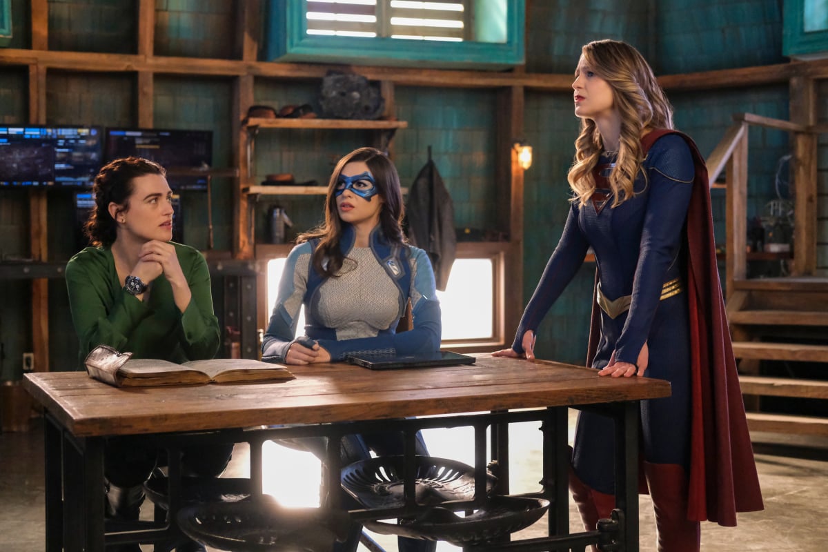 'Supergirl' Season 6 Episode 14 actors Katie McGrath, Nicole Maines, and Melissa Benoist, in character as Lena Luthor, Dreamer, and Supergirl, gather around a table. Dreamer and Supergirl are in their superhero costumes and Lena wears a green long-sleeved shirt.