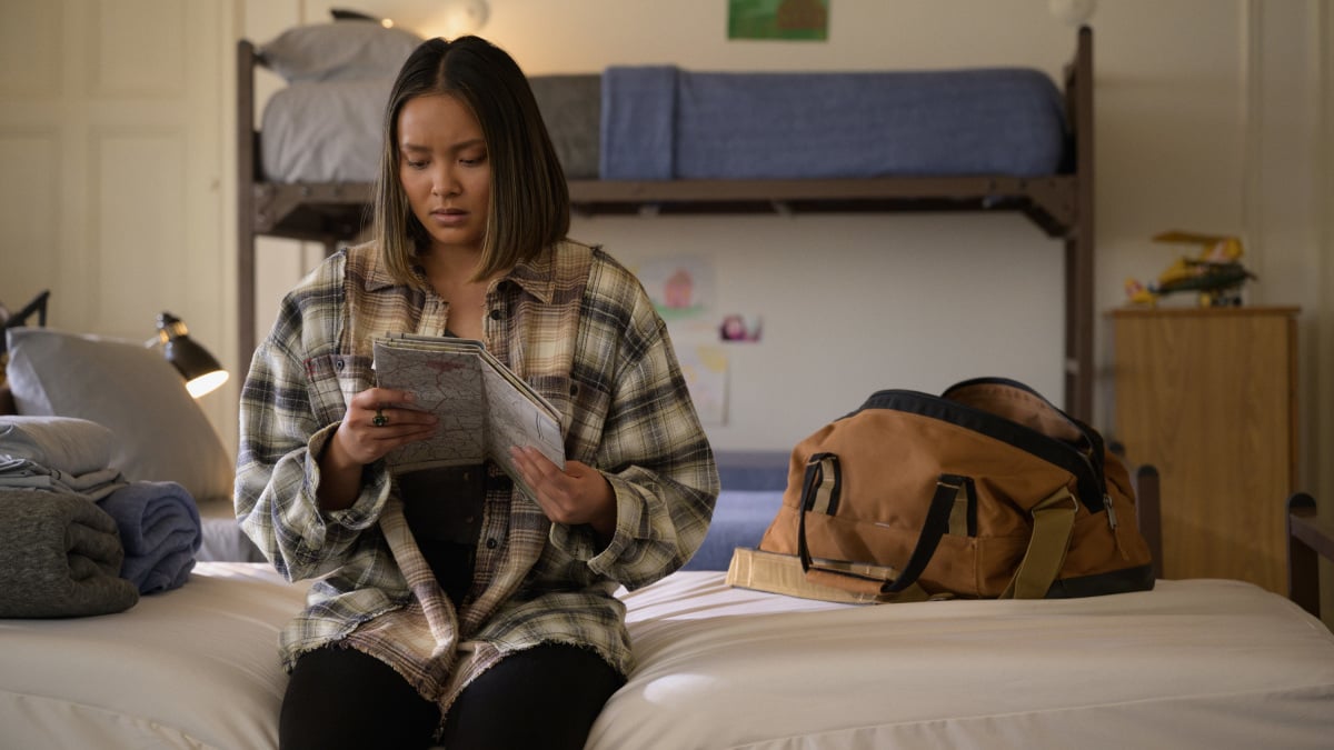 'Stargirl' Season 2 actor Ysa Penarejo, in character as Green Lantern's daughter Jennie, wears a plaid shirt and dark pants and sits on a bed looking at a map.