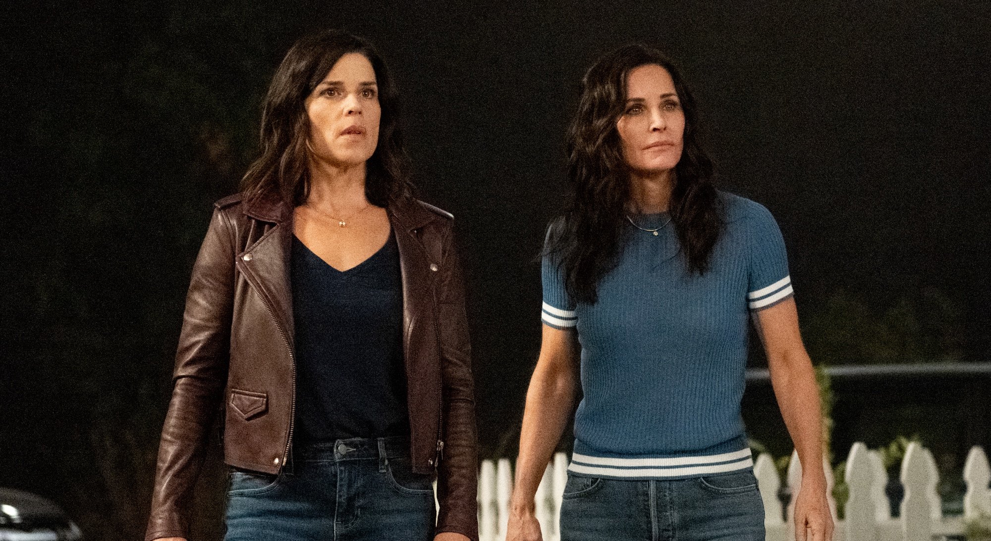 'Scream 5' cast members Neve Campbell as Sydney Prescott and Courteney Cox as Gale Weathers standing in front of a white fence looking shocked
