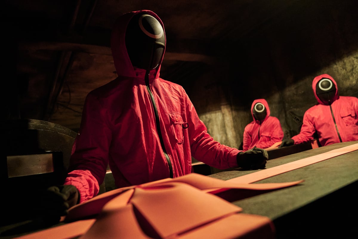 Workers in a pink and black jumpsuits move black coffins tied with pink bows in Squid Game.