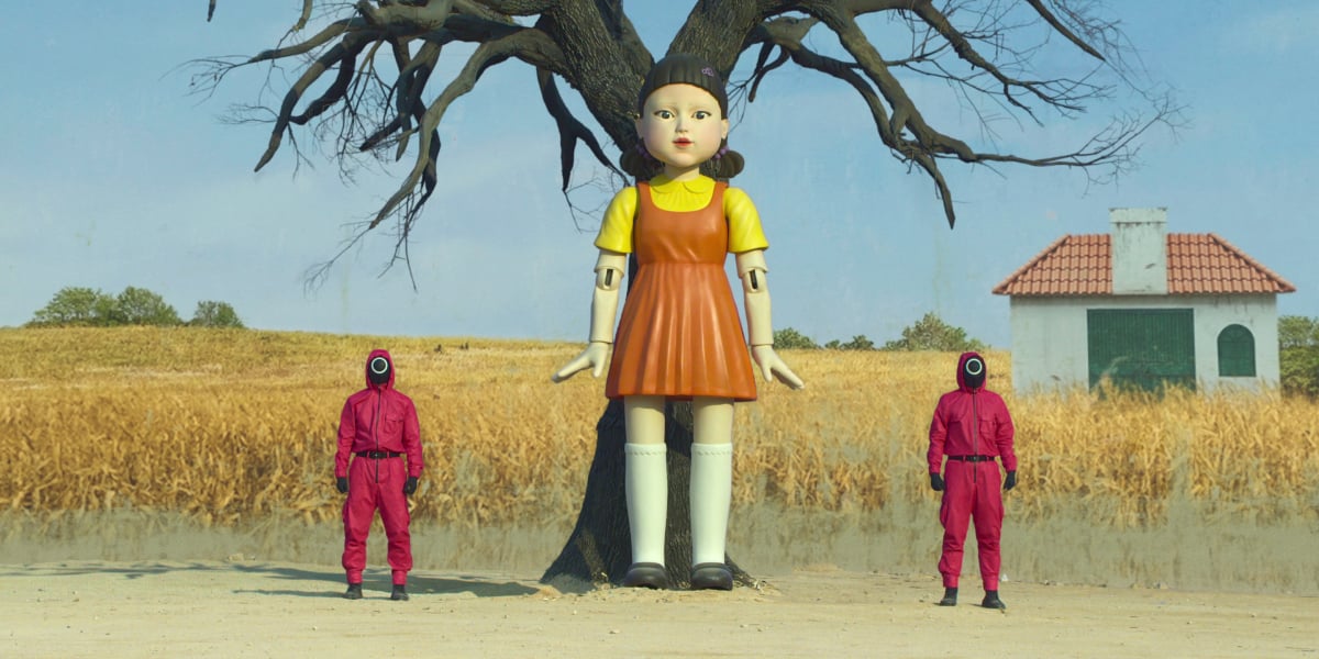 Two workers in pink jumpsuits stand beside the 'Squid Game' doll in episode 1.