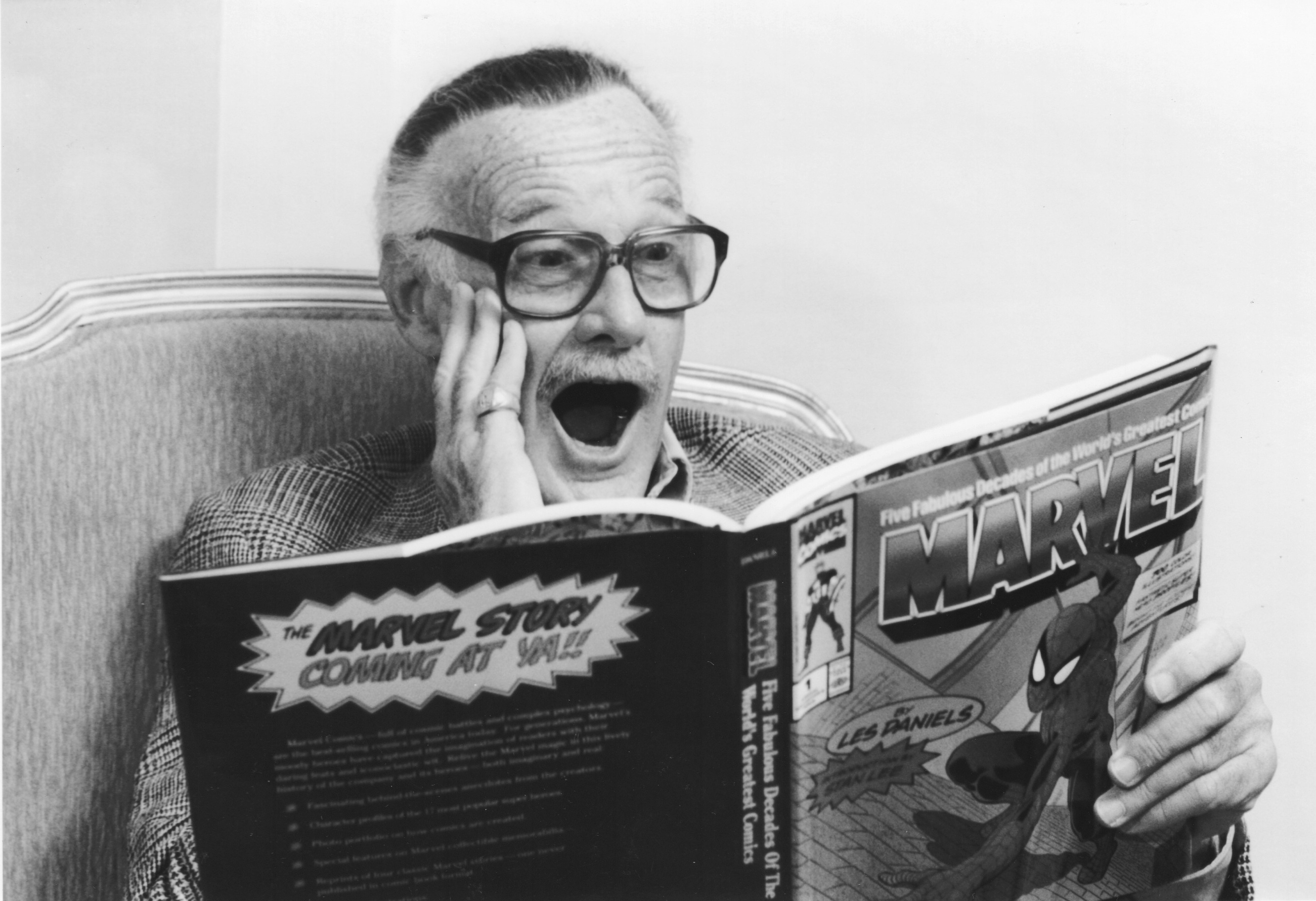 Marvel Comics legend Stan Lee reads a Marvel comic book. His hand is placed on his cheek and his mouth is agape. He is wearing glasses. The photo is in black and white.
