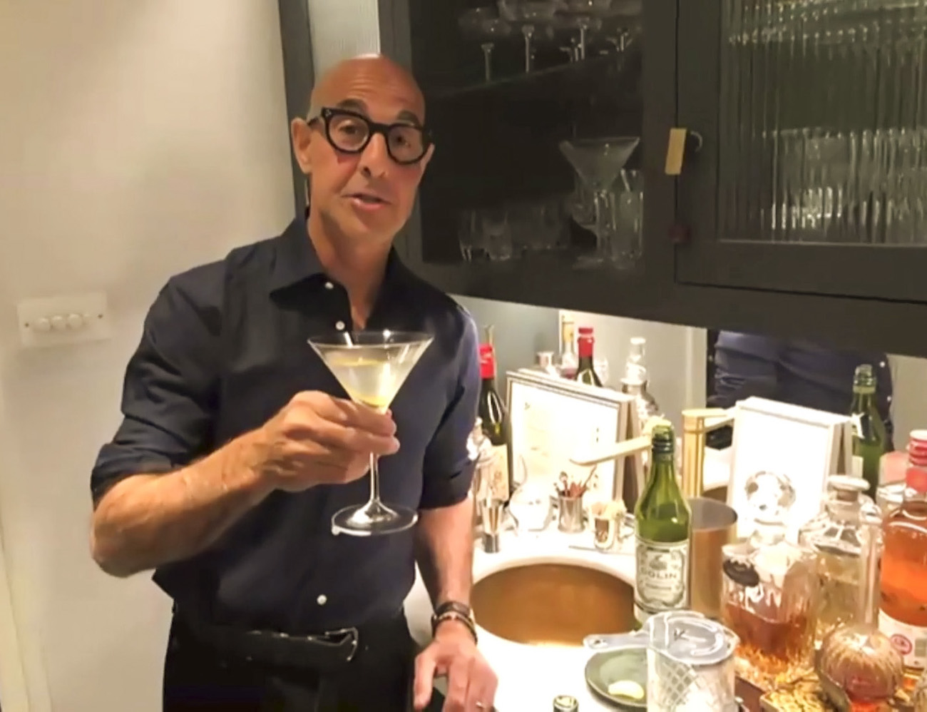 Stanley Tucci holds up a martini glass wearing a navy button-down shirt