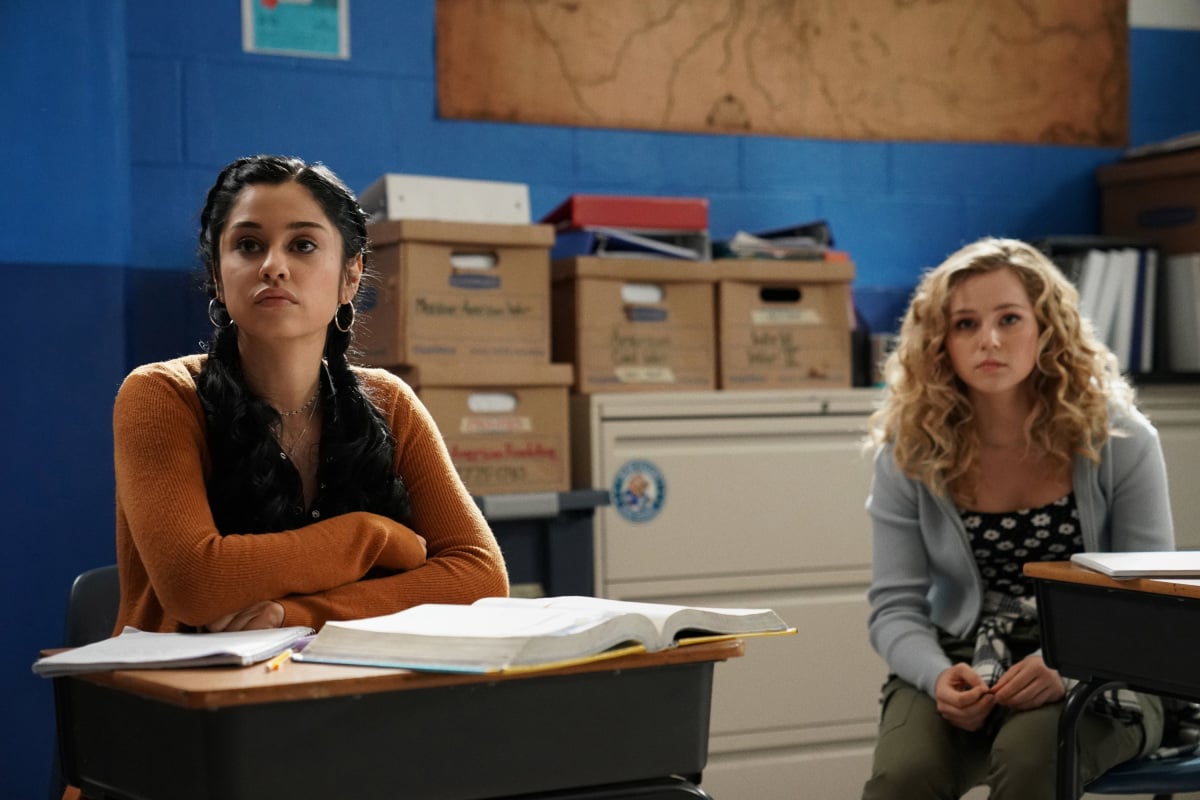 'Stargirl' actors Yvette Monreal and Brec Bassinger, in character, sit at desks in a classroom. Monreal wears a brown cardigan. Bassinger wears a gray cardigan over a black shirt with daisies. A sneak peek of 'Stargirl' Season 2 was shown at DC FanDome 2021.