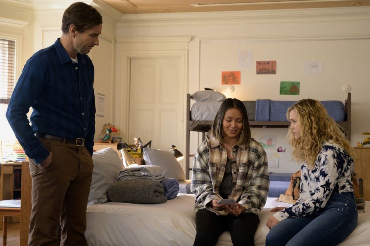 'Stargirl' actors Luke Wilson, Ysa Penarejo, and Brec Bassinger, all in character as Pat, Jennie, and Courtney, gather in a youth rehabilitation center. Jennie and Courtney sit on a bed while Pat stands above them. Pat wears brown pants and a blue button-up shirt. Jennie wears a white and green plaid shirt and black pants. Courtney wears a white and black tie dye hoodie and jeans.