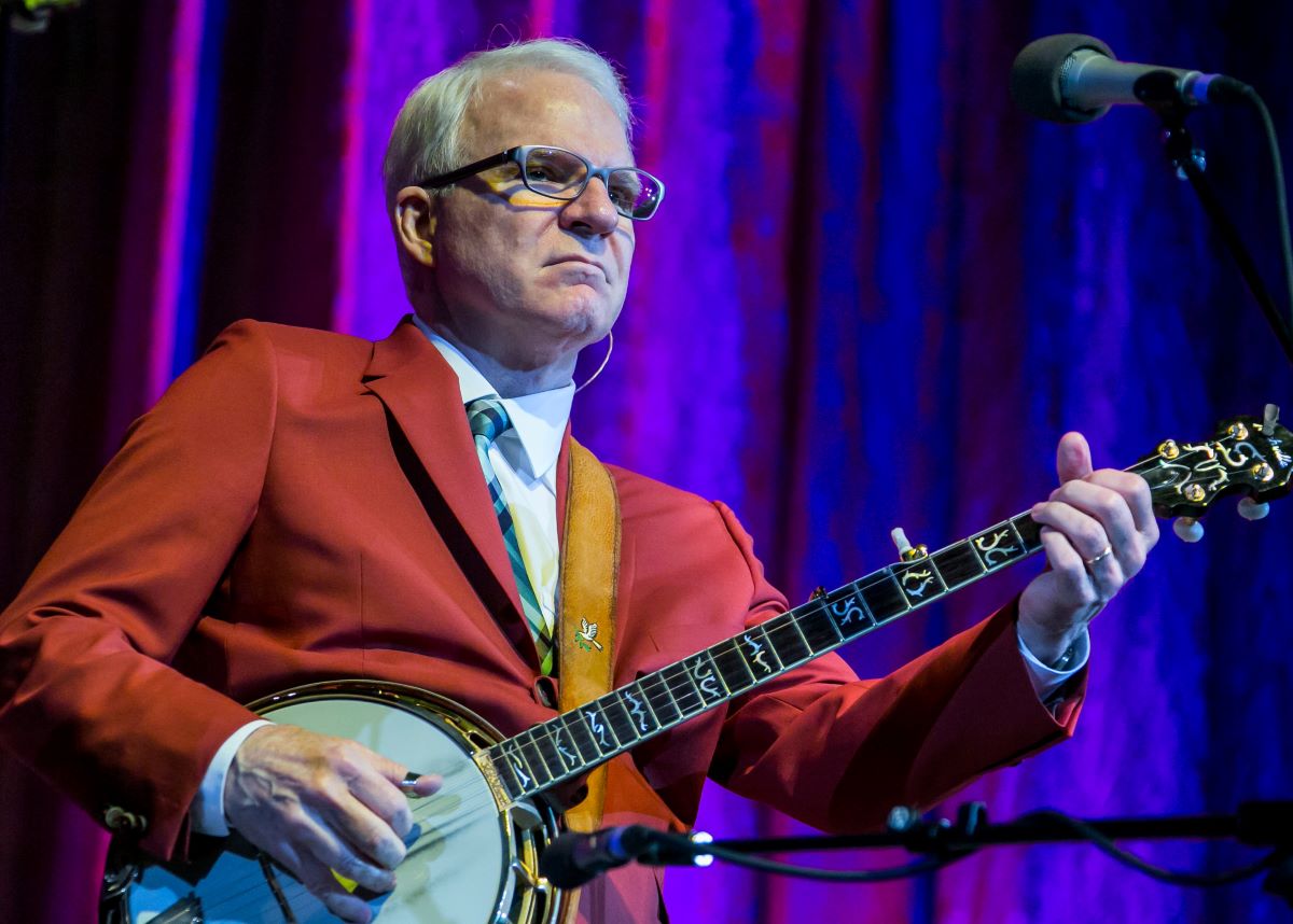 Steve Martin plays his banjo in a red jacket.