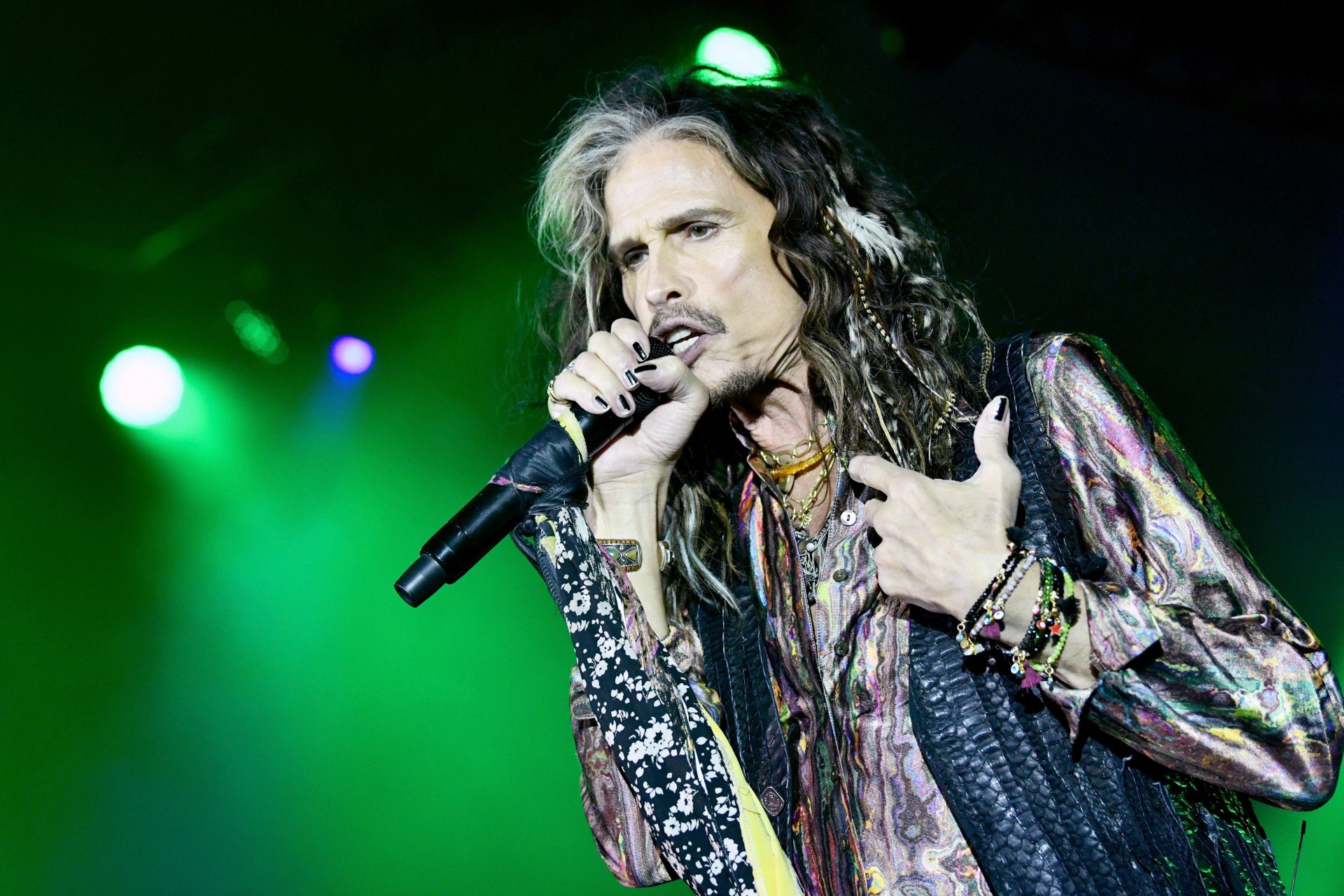 Aerosmith's Steven Tyler performs while holding a microphone and wearing a leather vest. 