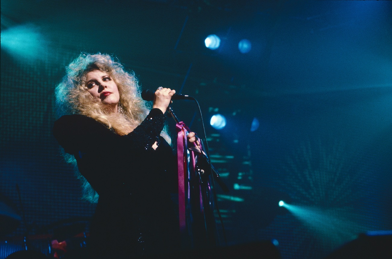 Stevie Nicks wears a black shirt and holds a microphone tied with pink ribbons. 