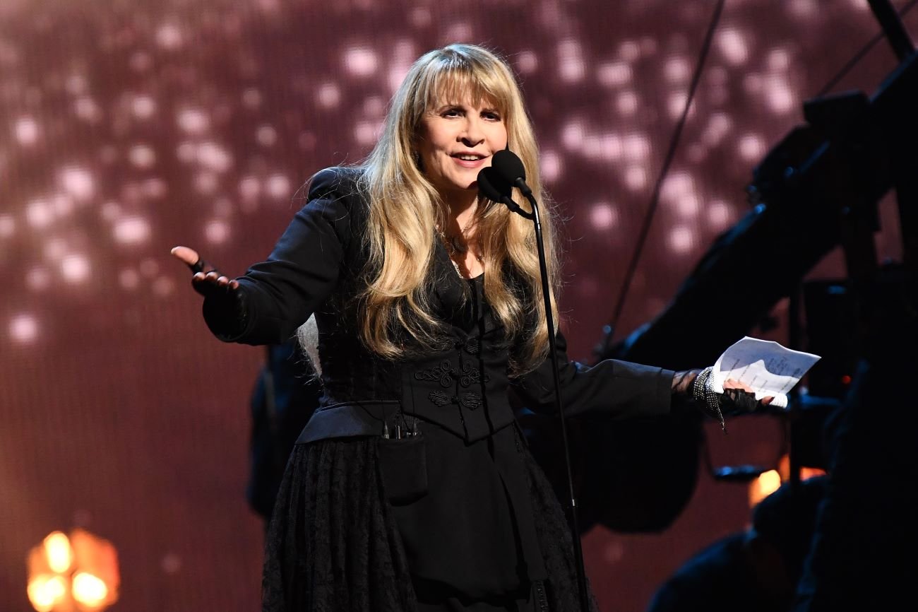 Stevie Nicks wears a black dress and holds a piece of paper. She stands in front of a purple background. 