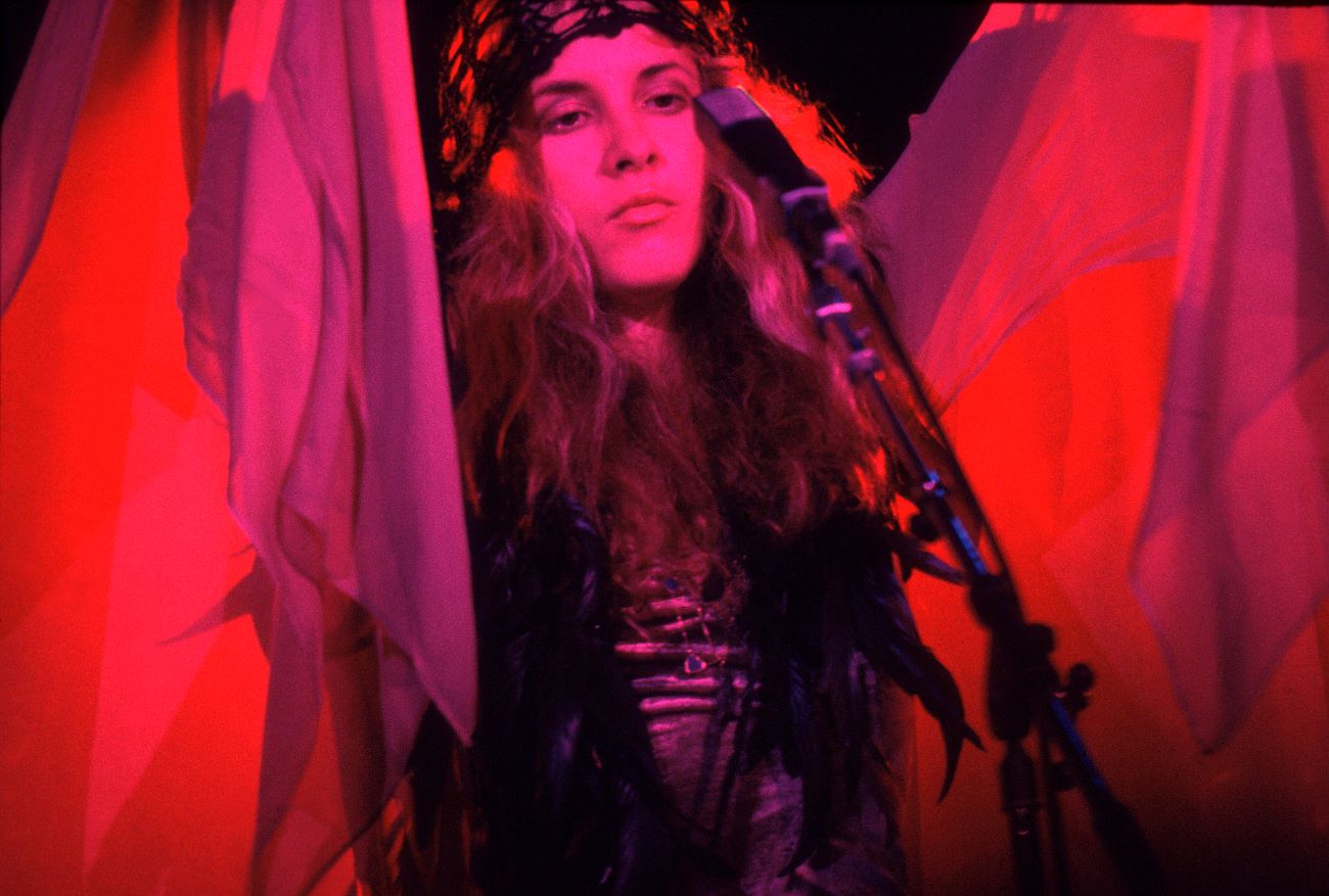 Stevie Nicks wears a head band and a shawl in pink lighting. She stands in front of a microphone. 