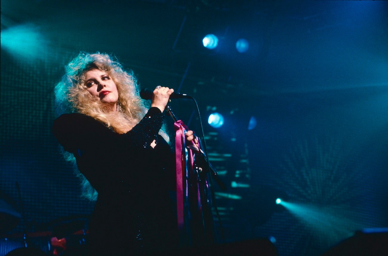 Stevie Nicks wears a black top and sings at a microphone with a pink ribbon tied around it while performing with Fleetwood Mac.