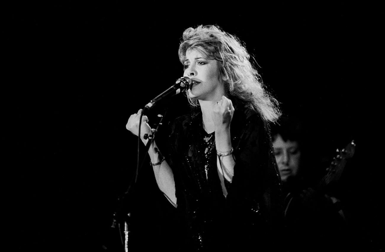 Stevie Nicks wears a black dress and sings into a microphone against a black background. 
