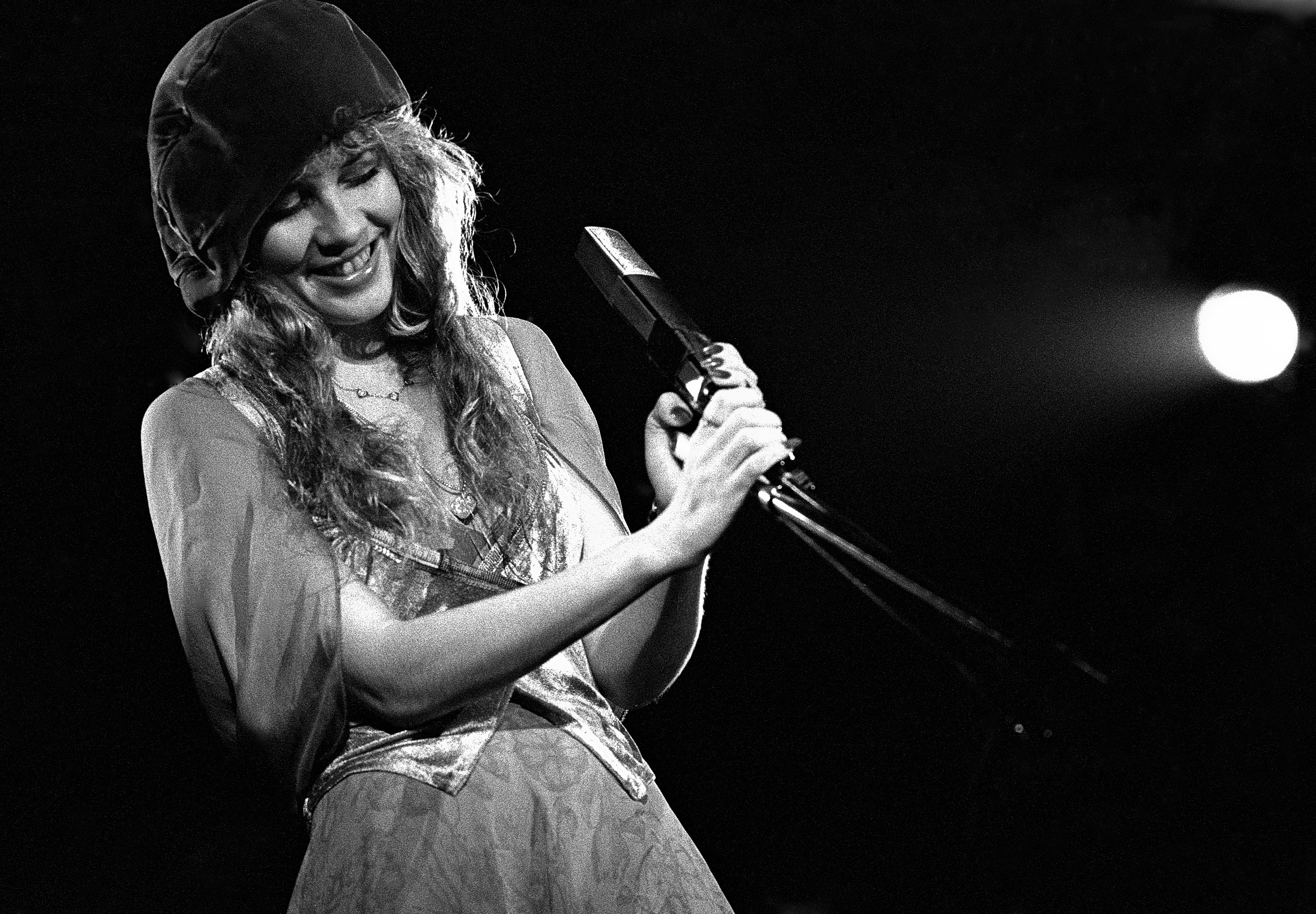A black and white photo of Stevie Nicks wearing a dress and a hat. She holds a microphone.