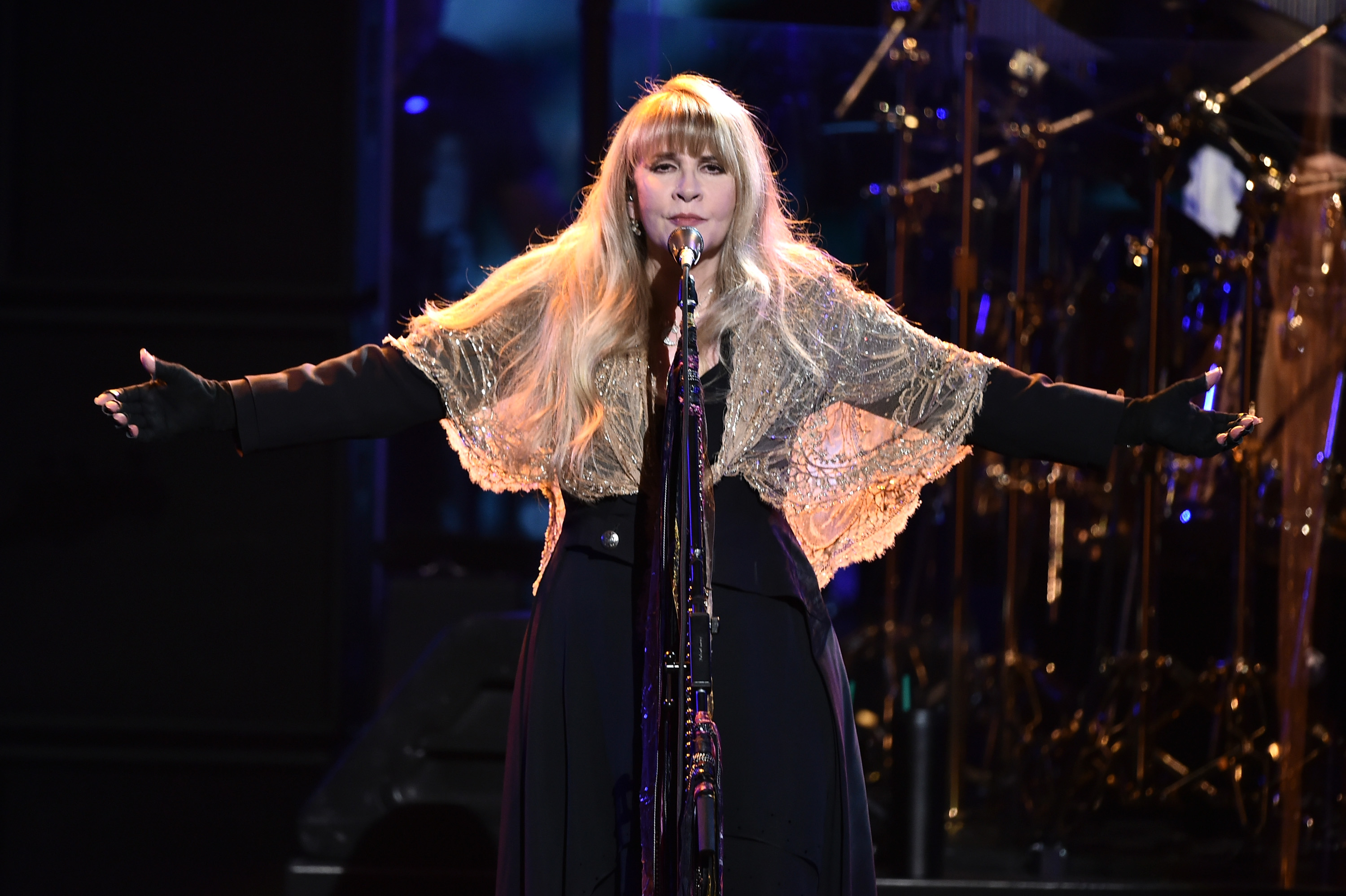 Stevie Nicks wears a black dress and sheer shawl. She stands in front of a microphone with her arms held wide.