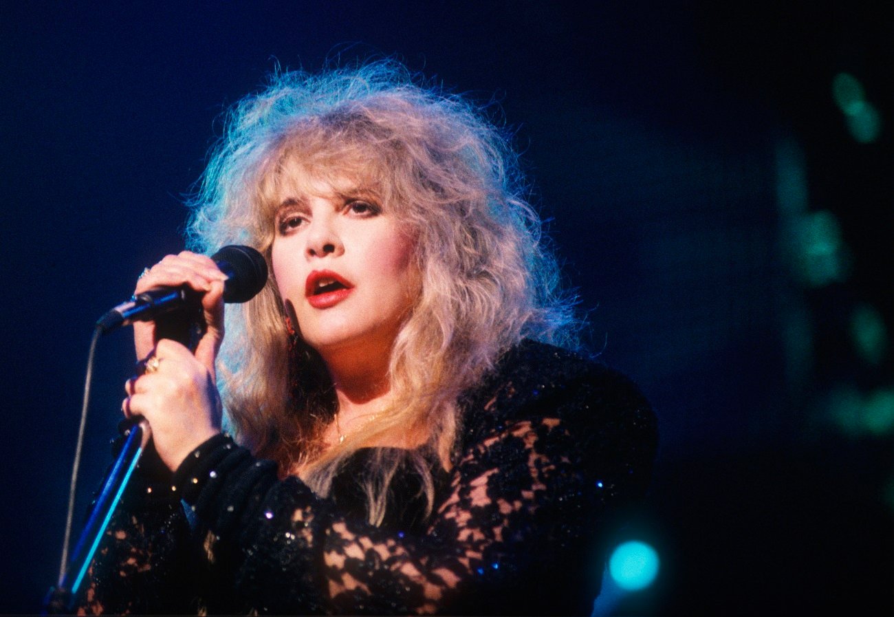 Stevie Nicks wears a black lace top and red lipstick. She holds a microphone.