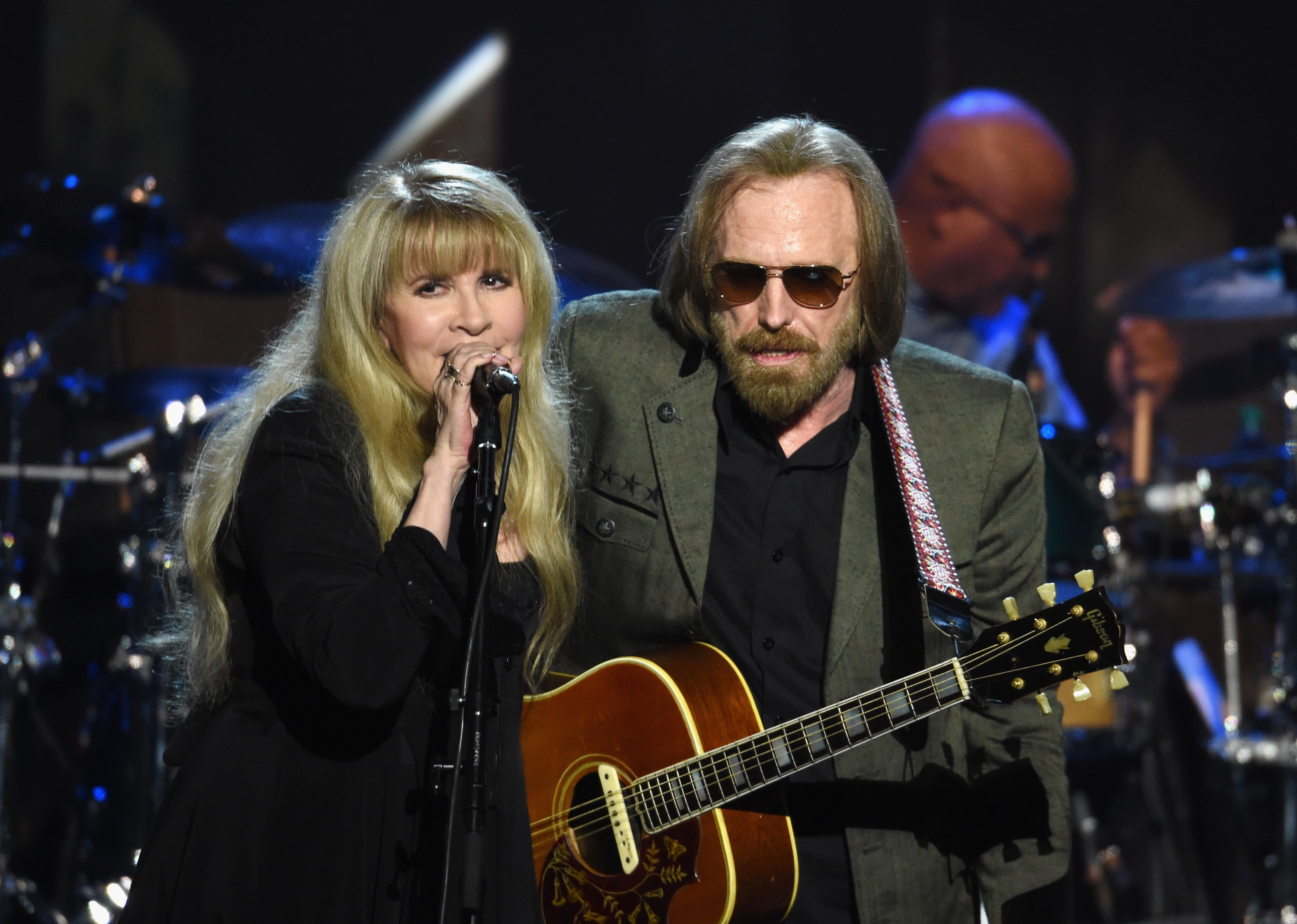 Stevie Nicks in a black dress and Tom Petty in a gray jacket with a guitar.