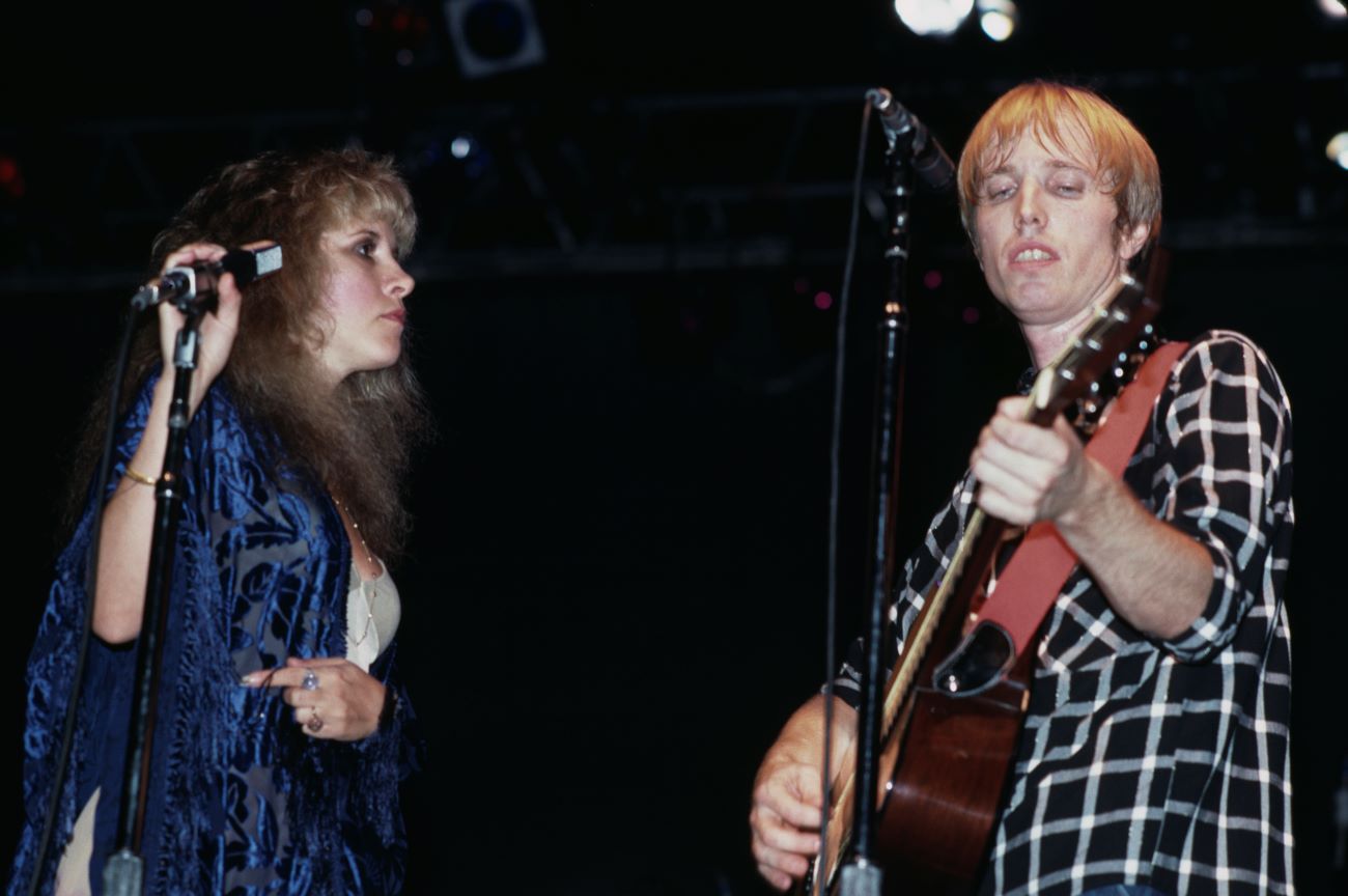 Stevie Nicks and Tom Petty on stage together. She wears a blue shawl and he wears a black plaid shirt.