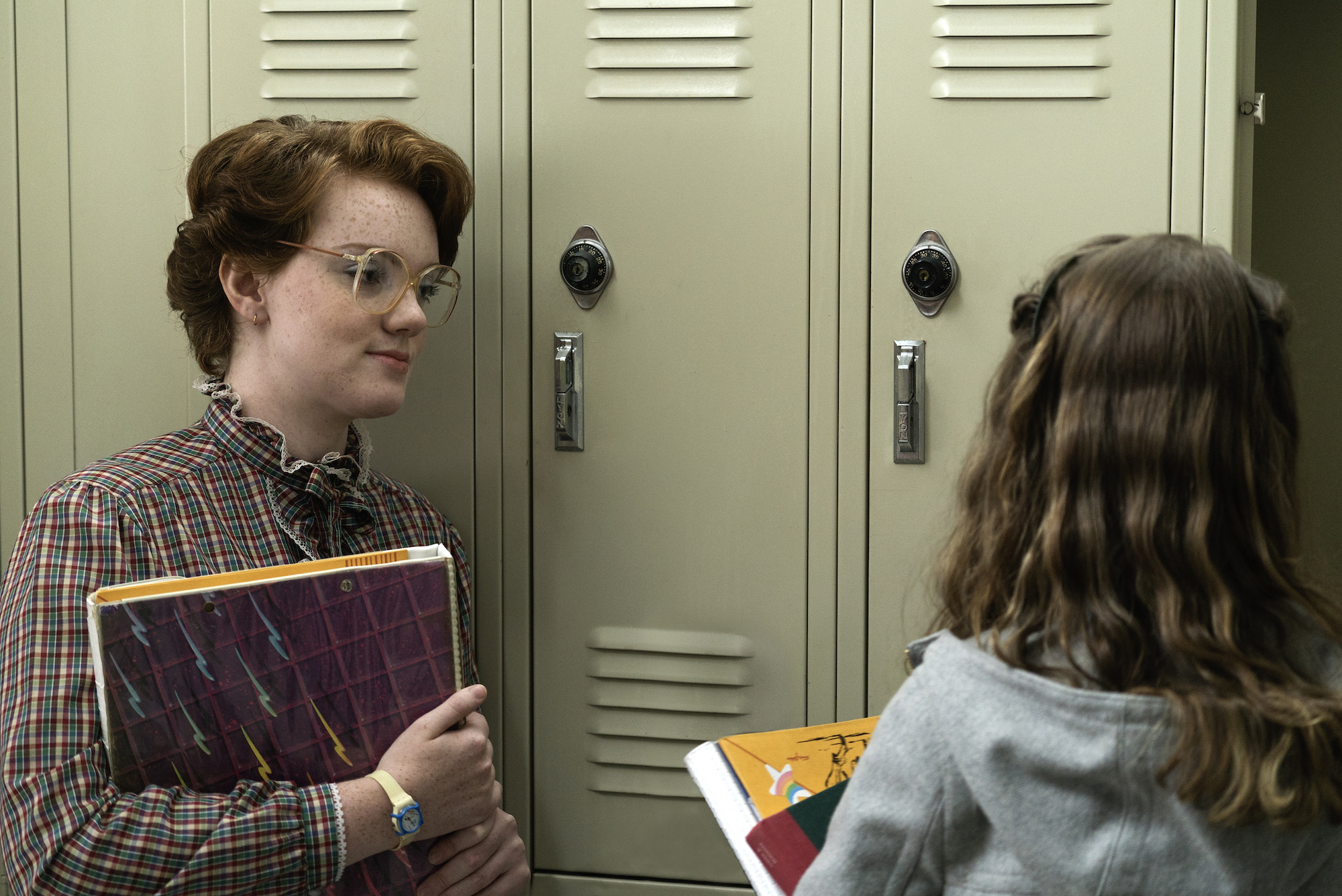 Stranger Things character Barb was one of the first deaths of season 1. She's seen here in a production still leaning against some lockers talking to Nancy Wheeler.