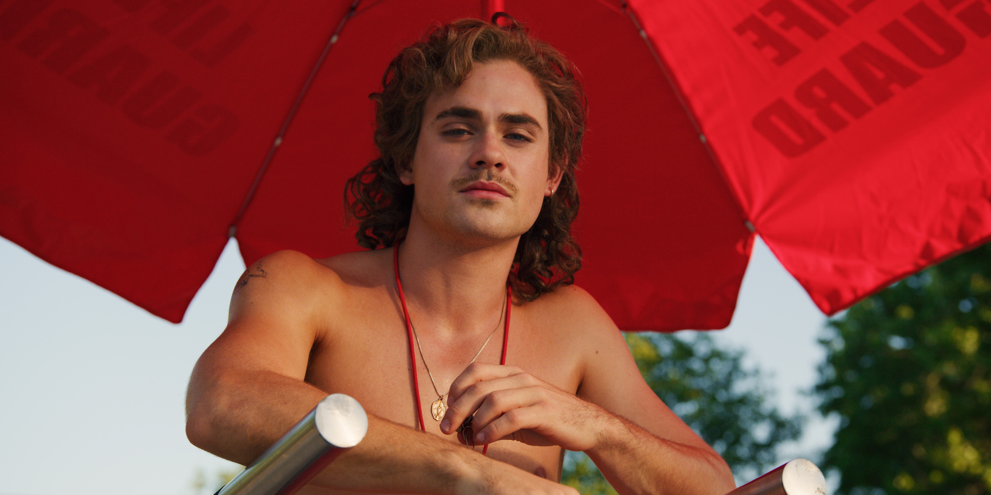 Dacre Montgomery as Billy Hargrove sits in a lifeguard chair in a production still from 'Stranger Things' Season 3.