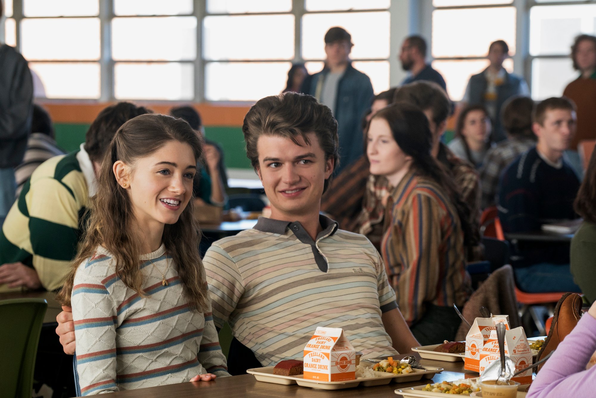 Nancy and Steve smile in the cafeteria at their high school in a production still from 'Stranger Things' Season 1.