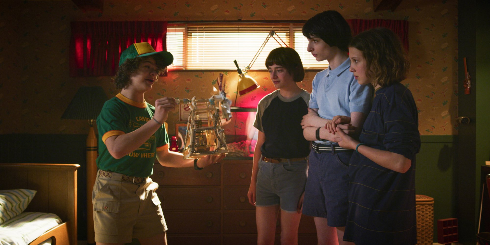 Dustin holding out a toy to Will, Mike, and Eleven in a production still from 'Stranger Things' Season 3