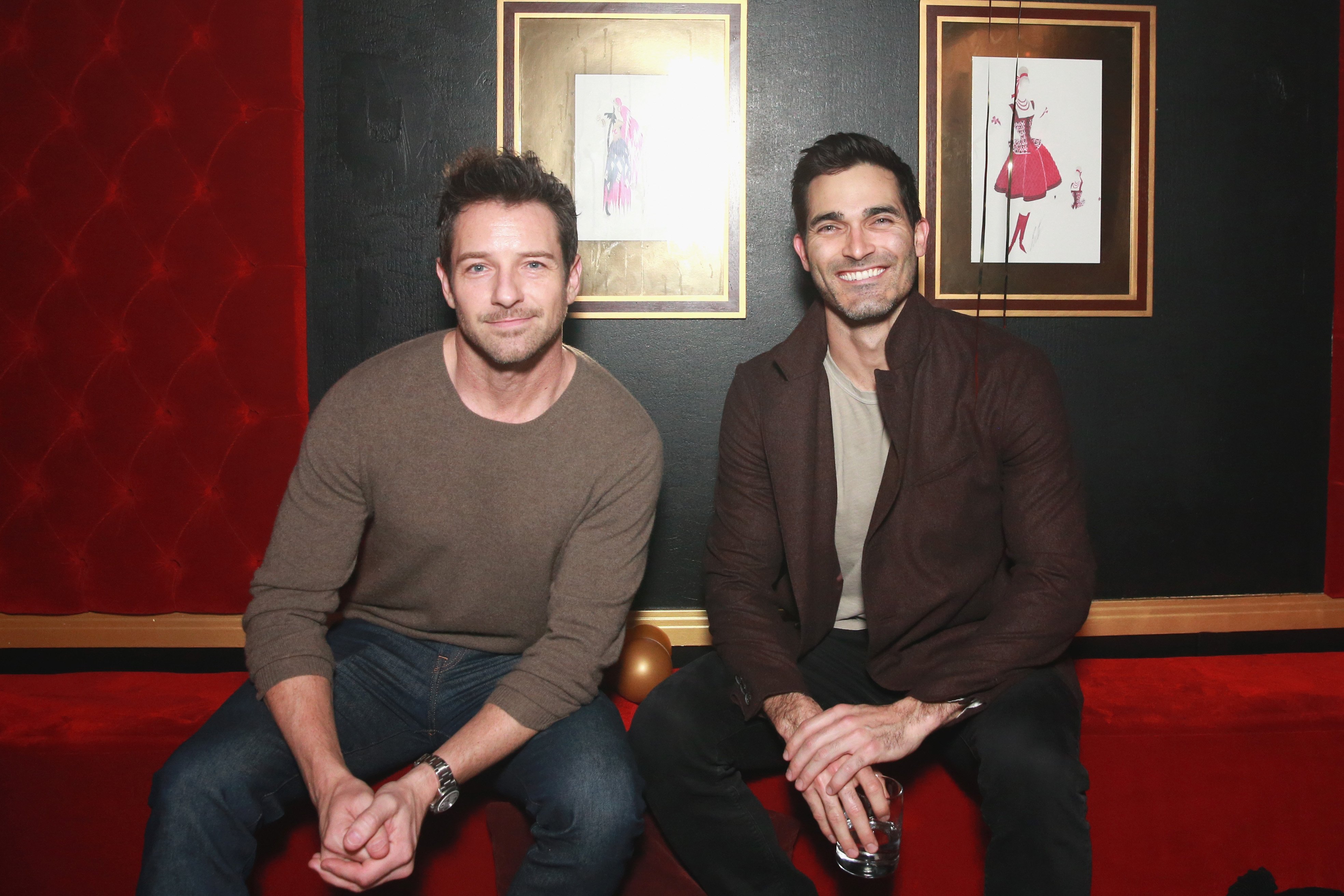 'Superman & Lois' and 'Teen Wolf' co-stars Ian Bohen and Tyler Hoechlin sit together on a red coach. Bohen wears a tan sweater and jeans. Hoechlin wears a dark brown jacket over a gray shirt and black pants.