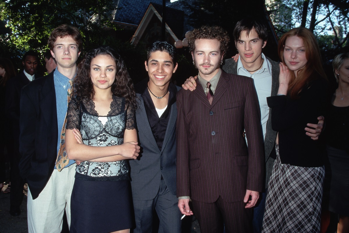 'That '70s Show' actor Topher Grace in khakis, blue shirt and navy blazer, Mila Kunis is a black and white dress, Wilmer Valderrama in a grey suit and black shirt, Danny Masterson in a brown suit, Ashton Kutcher in a grey suit, and Laura Prepon in a black blouse and plaid skirt, pose for a cast photo.
