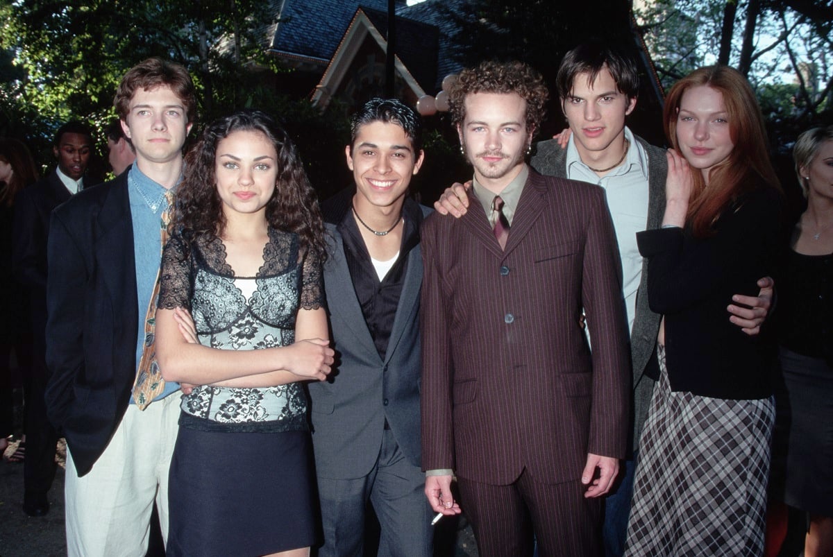 'That '70s Show' actor Topher Grace in khakis, blue shirt and navy blazer, Mila Kunis is a black and white dress, Wilmer Valderrama in a grey suit and black shirt, Danny Masterson in a brown suit, Ashton Kutcher in a grey suit, and Laura Prepon in a black blouse and plaid skirt, pose for a cast photo.