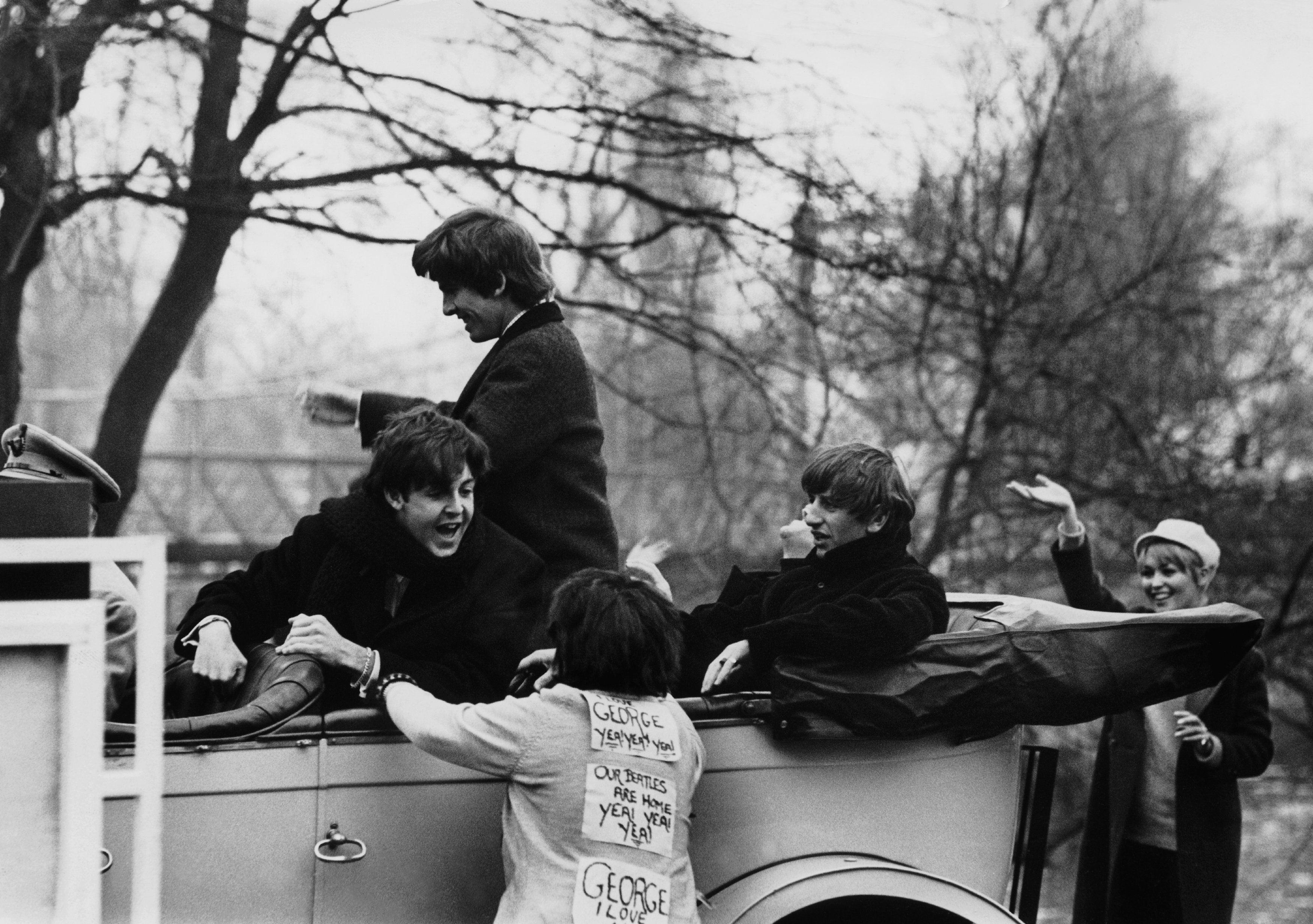 After their return from their tour in the U.S.A., The Beatles are cheered by two fans. They are in a convertible car driven by a chauffeur.