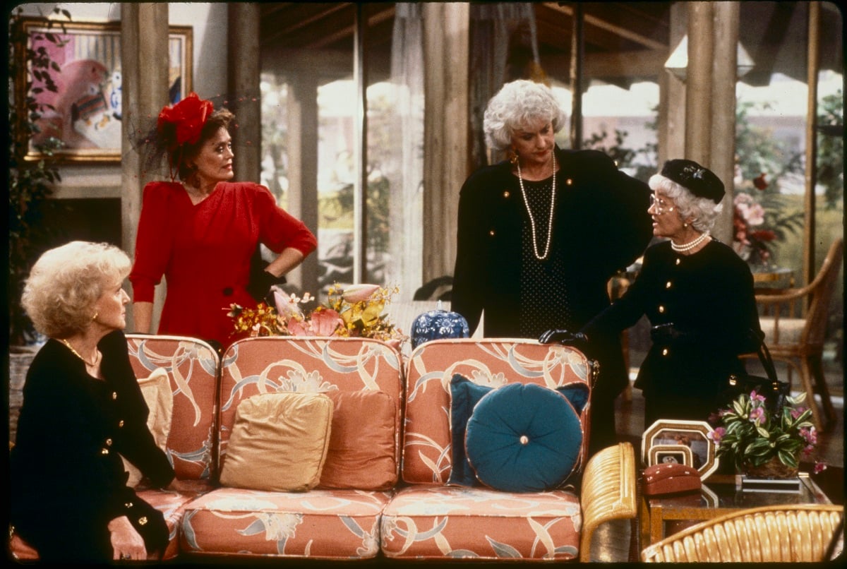Betty White in a black dress, Rue McClanahan in a red dress, Bea Arthur dressed in a black outfit, and Estelle Getty also wearing a black dress in a 'The Golden Girls' scene.