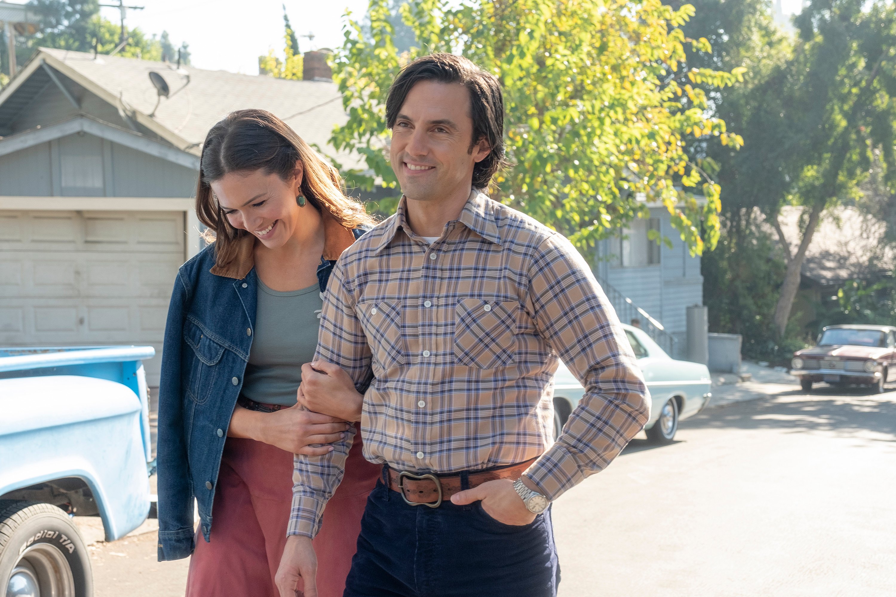 'This Is Us' Season 6 stars Mandy Moore and Milo Ventimiglia, in character as Rebecca and Jack, walk side-by-side, and Rebecca is holding onto Jack's arm. Rebecca is wears red pants, a green shirt, and a jean jacket. Jack is wearing a blue and tan long-sleeved plaid shirt and jeans.