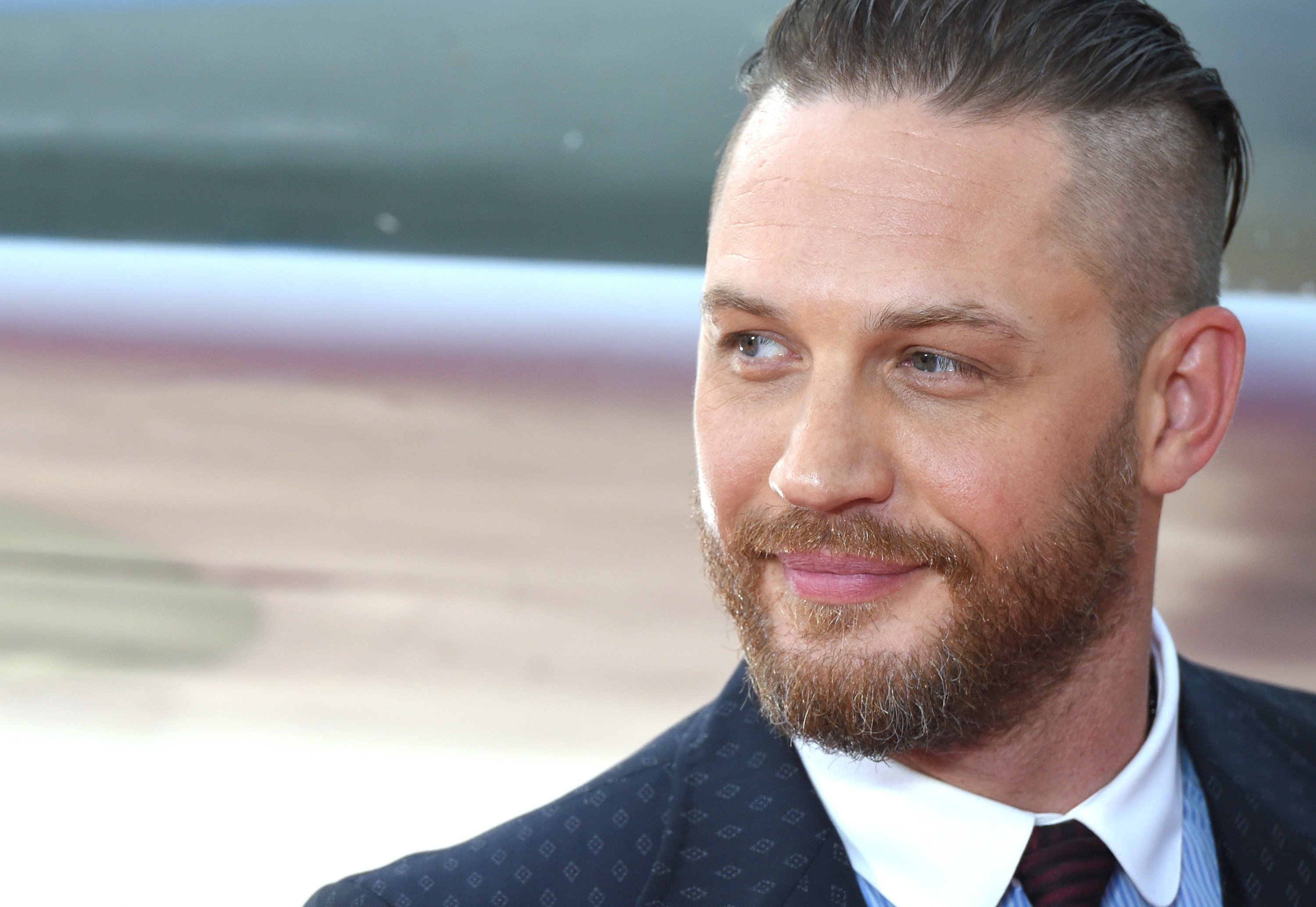 Tom Hardy, who plays Alfie in 'Peaky Blinders,' smiles for a photo wearing a suit and tie. He also has a beard.