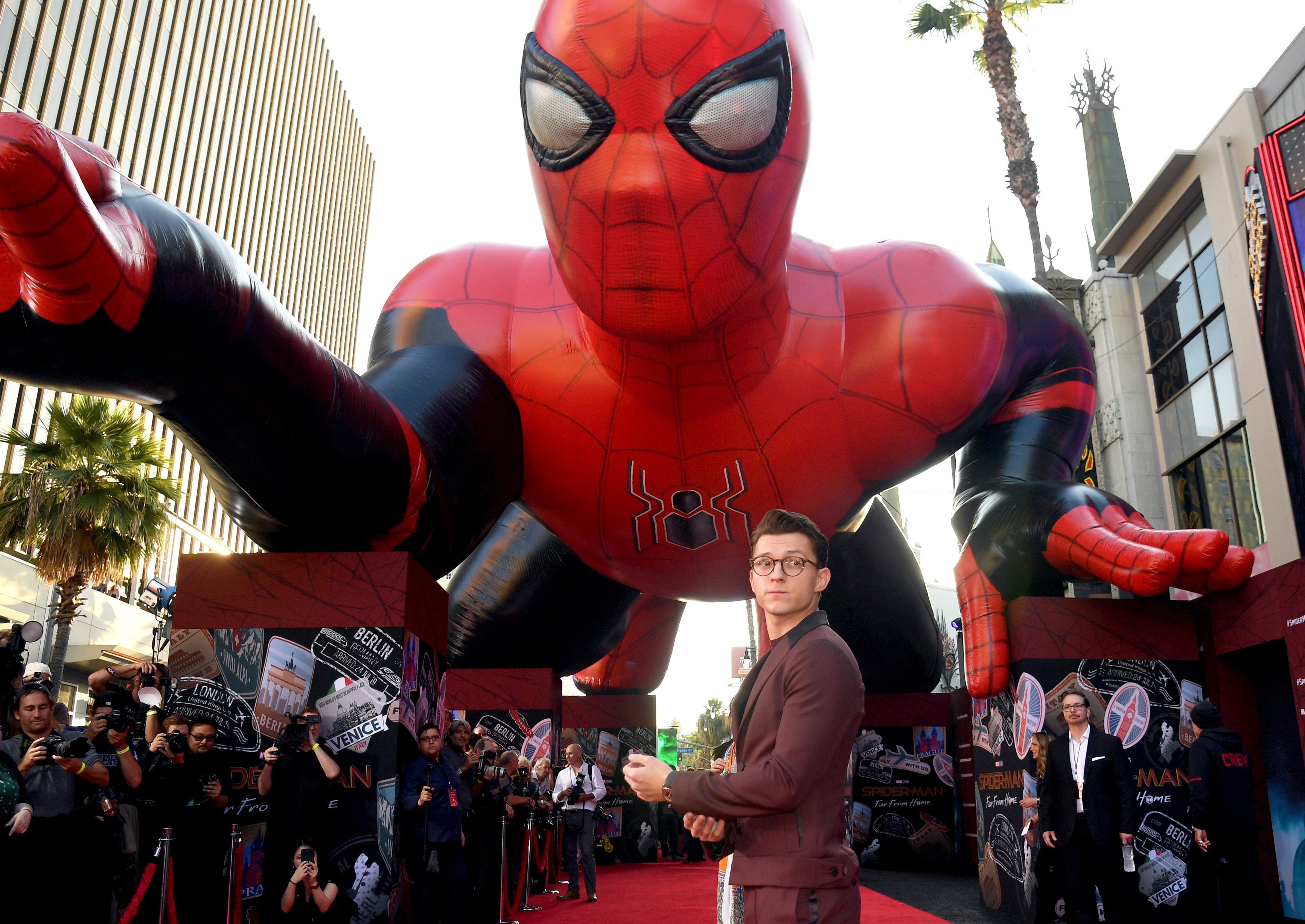 'Spider-Man: No Way Home' star Tom Holland walks the red carpet for a film premiere. He wears a maroon suit and glasses, and he stand in front of a huge blow-up Spider-Man balloon.