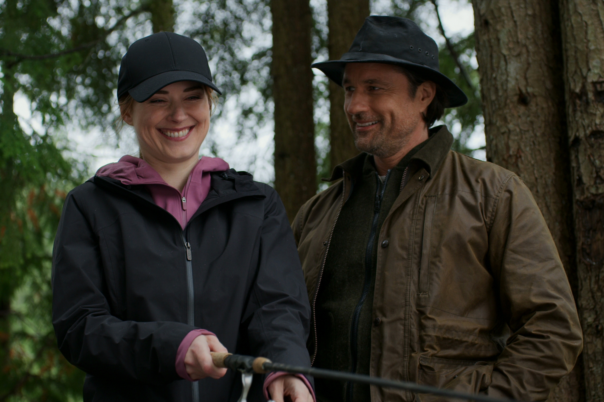 Alexandra Breckenridge as Melinda Monroe and Martin Henderson as Jack Sheridan in 'Virgin River' on Netflix. She's wearing a black hoodie and cap and smiling while holding a fishing rod. He's looking at her and smiling.