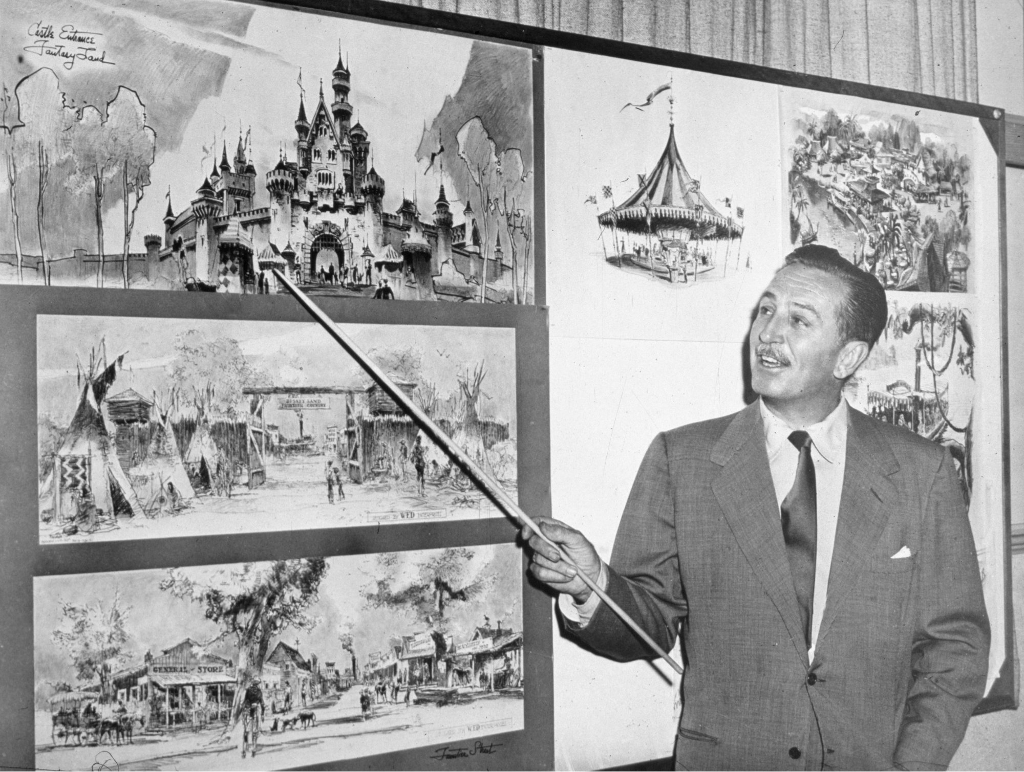 Walt Disney pointing at sketches of Disneyland with a pointer wearing a suit