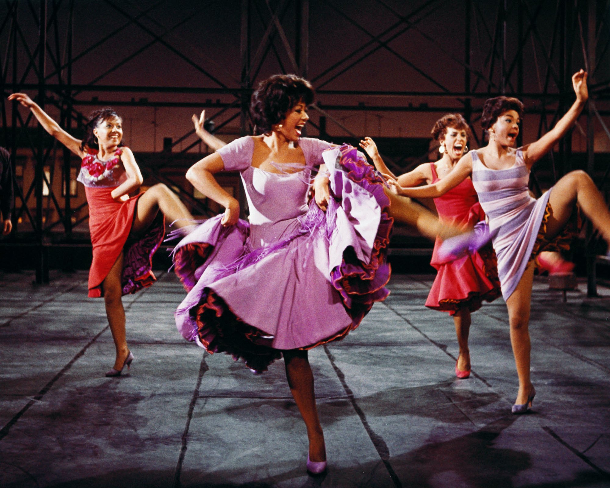 Rita Moreno performing 'America' in 'West Side Story' (1961). She wears her iconic purple dress as she dances with other female cast members on the New York City rooftop set at nighttime.