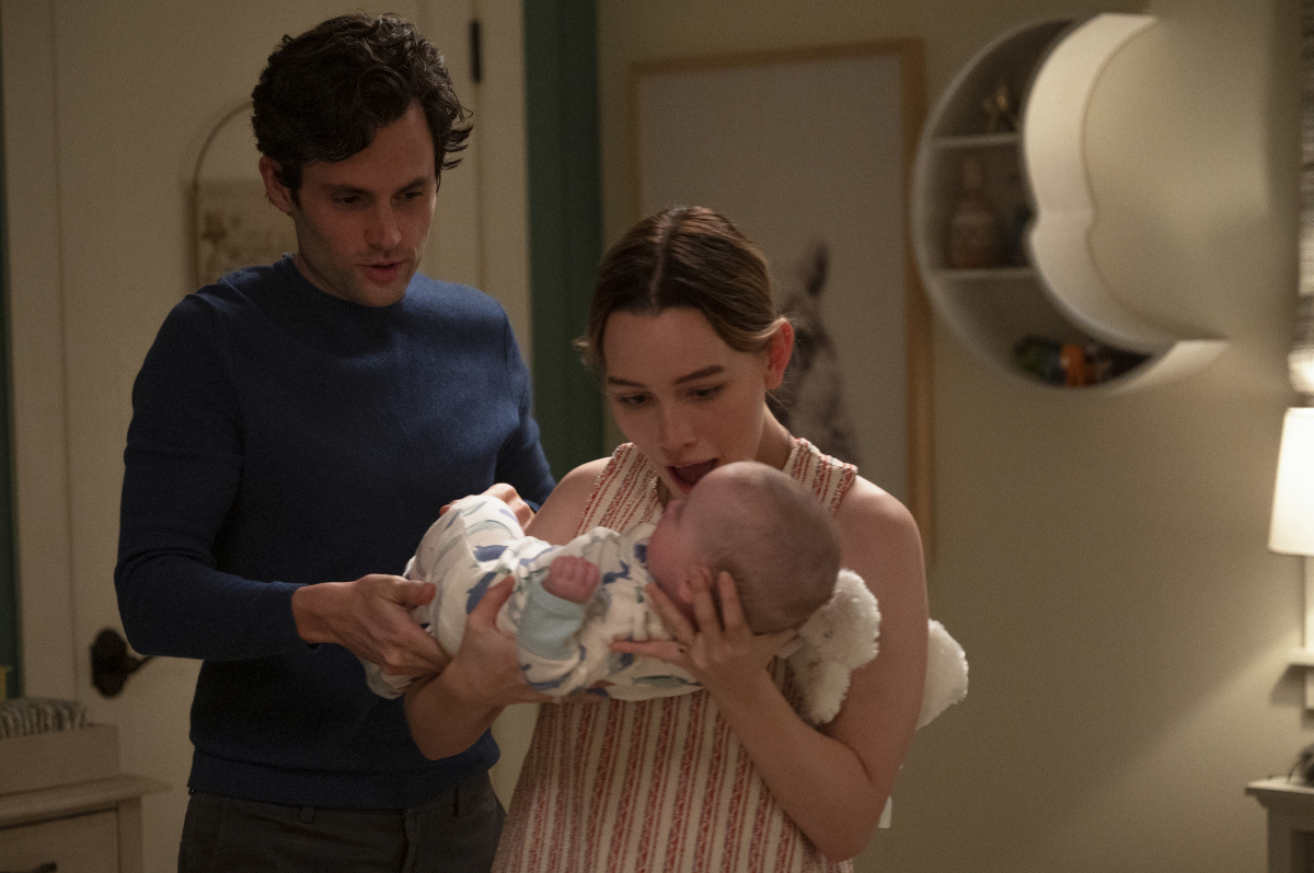 Love Quinn holds baby Henry while Joe Goldberg stands next to her in 'You' Season 3. Joe is wearing a blue sweater and Love is wearing a striped top.