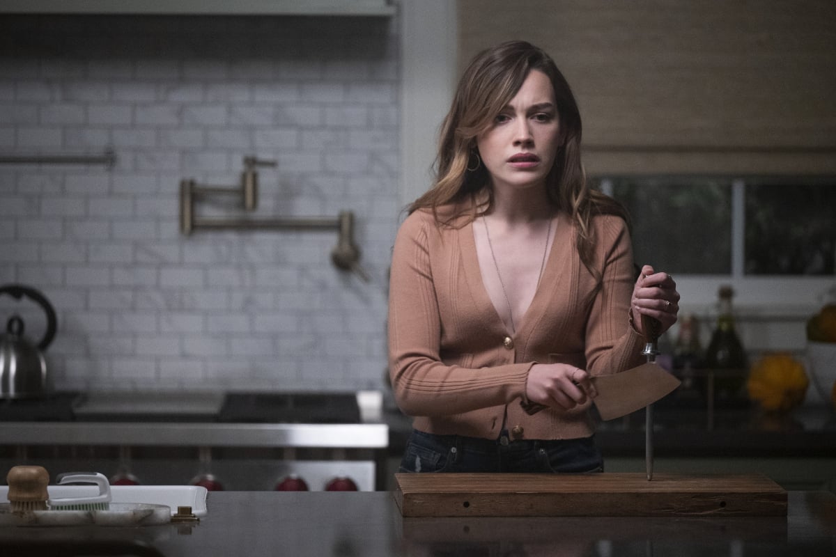Love Quinn stands in the kitchen sharpening a butcher's knife and looking upset in 'You' Season 3. Love is wearing a brown, low-cut top.