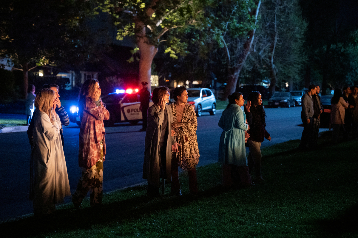 Surprised neighbors in You Season 3 wait outside their homes with police cars behind them.