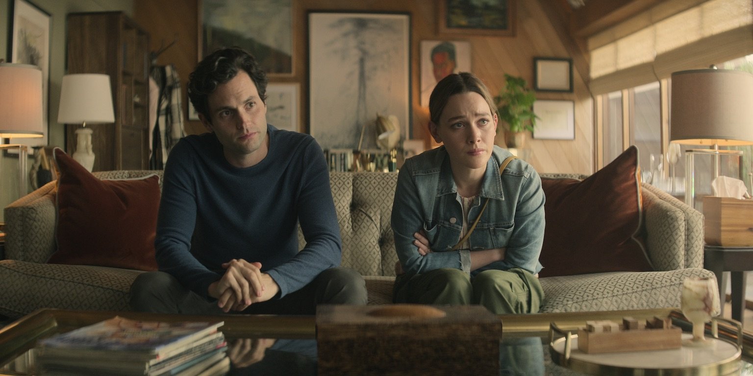 Penn Badgley and Victoria Pedretti from the 'You' Season 3 cast sitting next to each other on a couch looking worried