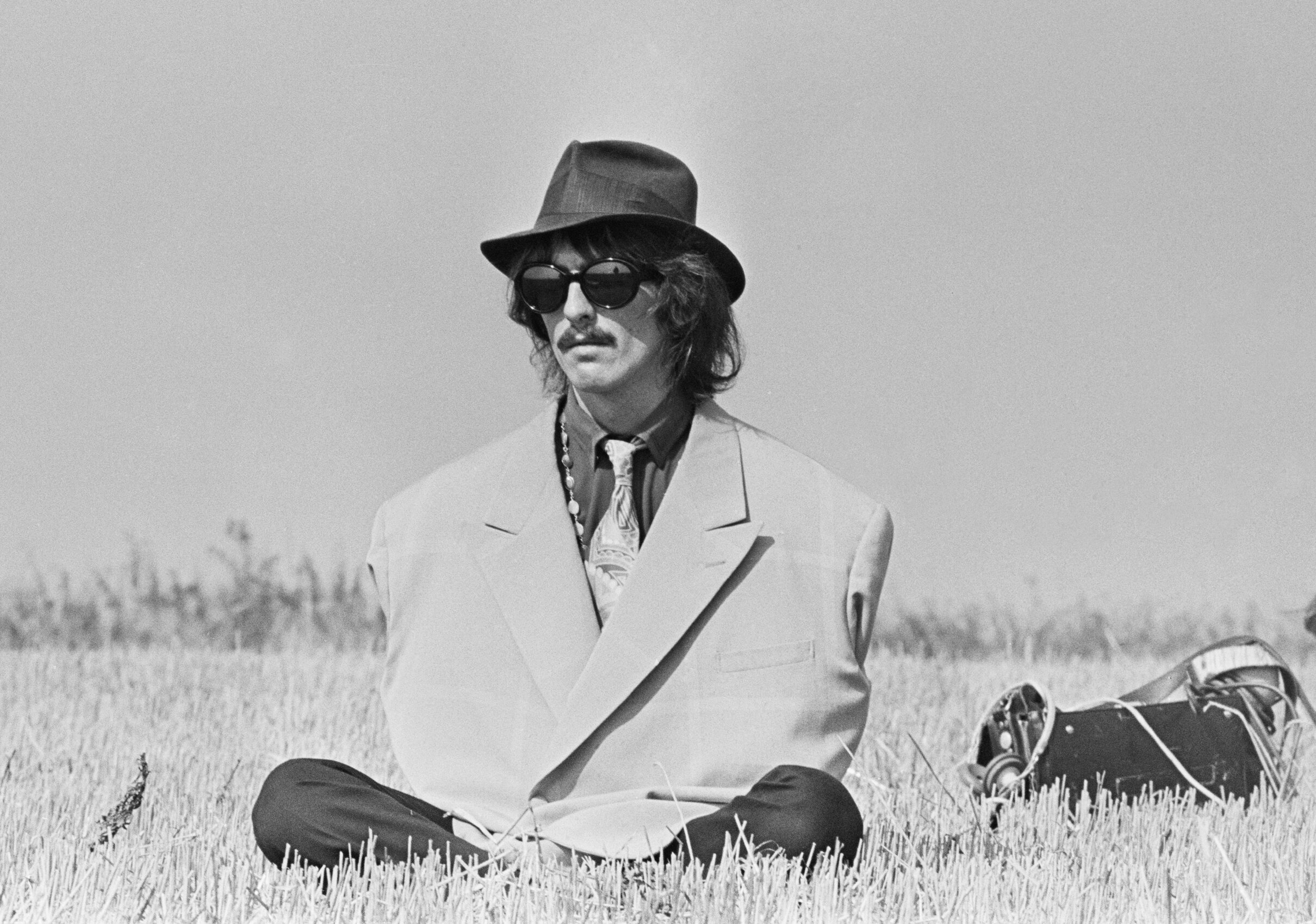 George Harrison sits on the ground with his arms inside his coat.