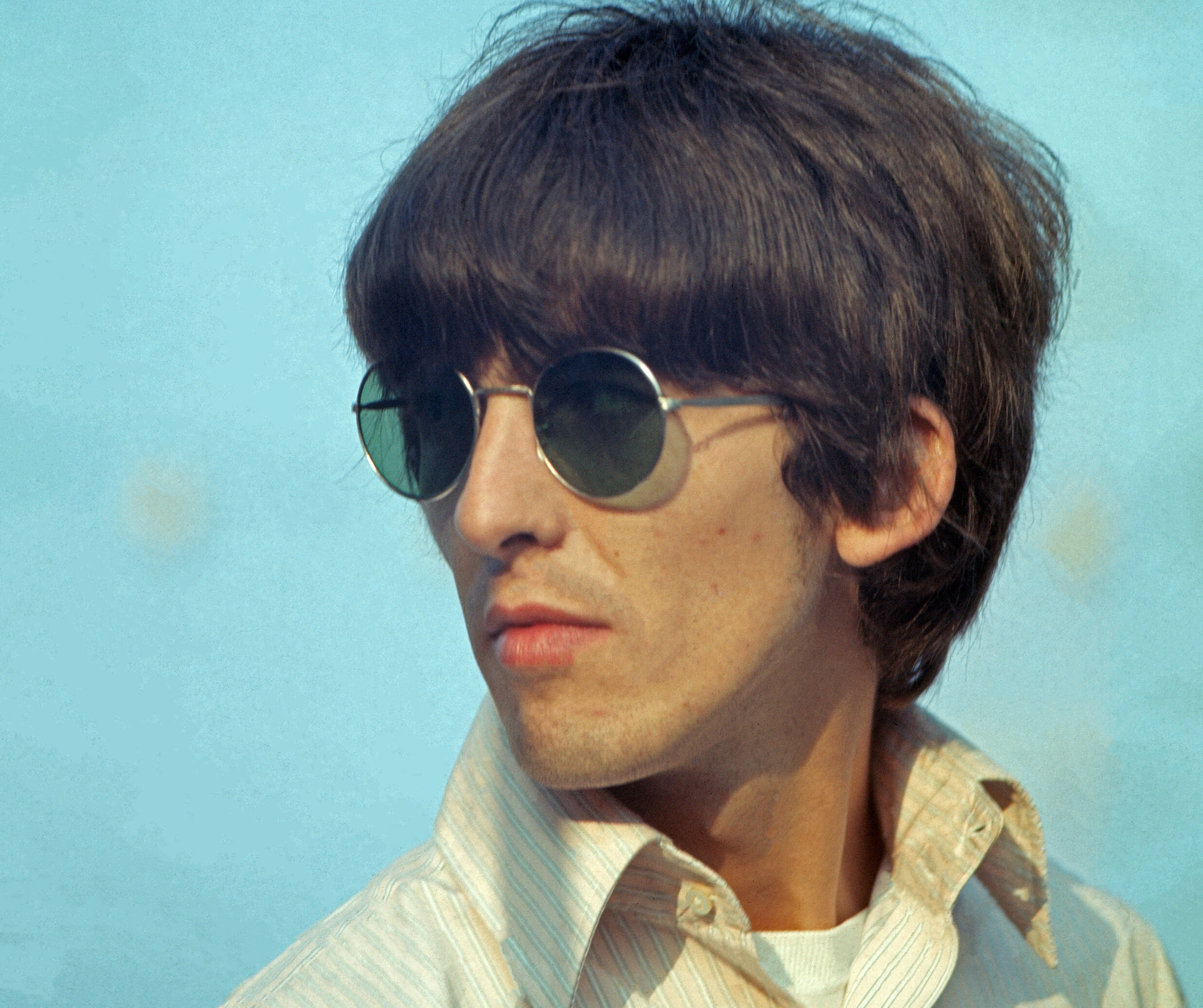 A close-up of George Harrison in 1966. He's wearing round sunglasses and a yellow collared shirt.