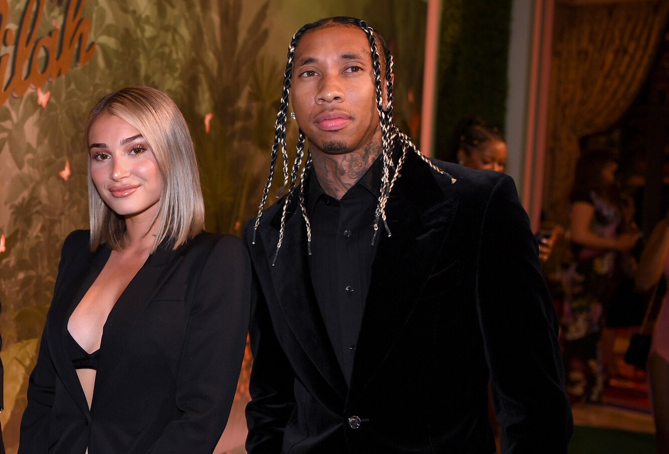 Camaryn Swanson smiling and wearing a black jacket stands next to Tyga, wearing a black jacket and black shirt.