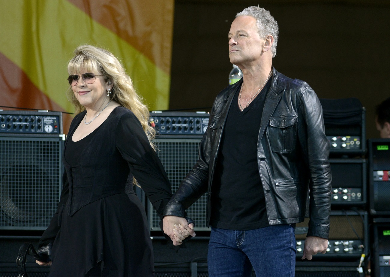Stevie Nicks wears a black dress and Lindsey Buckingham wears a black shirt and leather jacket. They hold hands onstage.