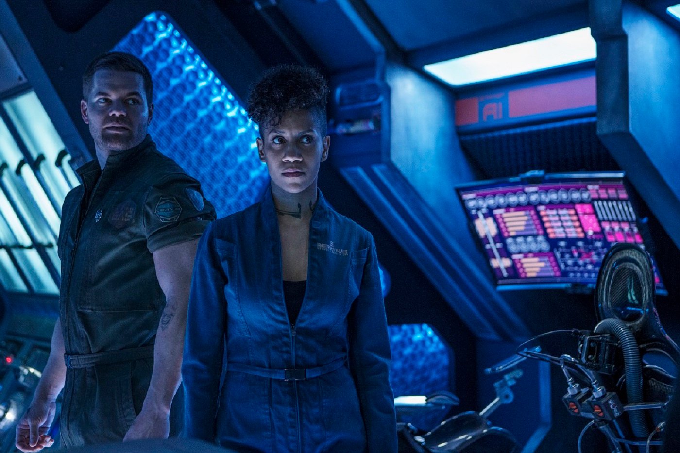 'The Expanse' stars Wes Chatham as Amos Burton and Dominique Tipper as Naomi Nagata -9 Amos wears a green jumpsuit, Naomi a blue one. They are aboard the Rocinante, looking serious, 