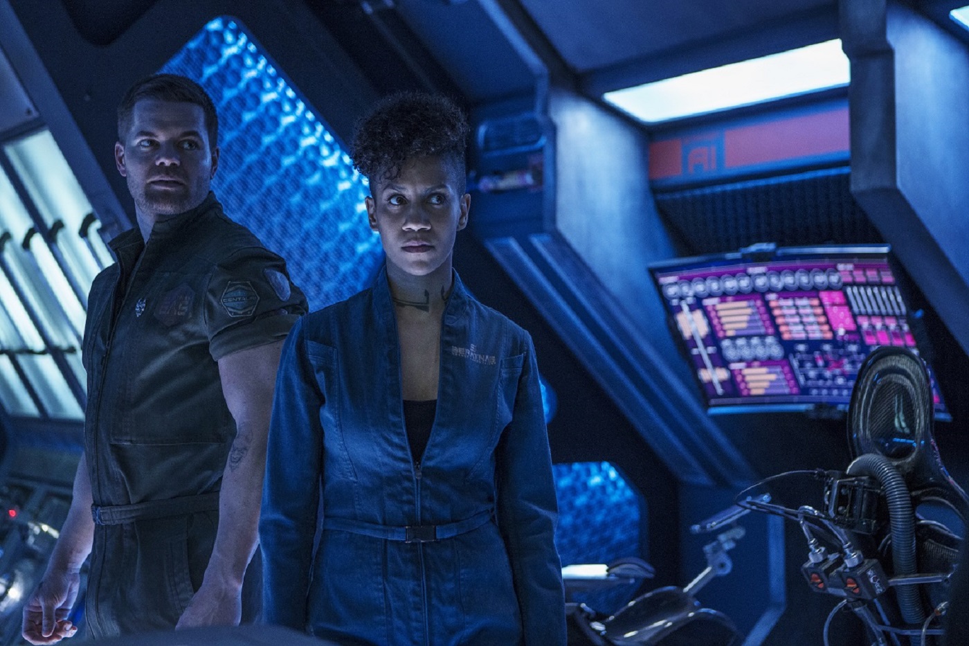 'The Expanse' stars Wes Chatham as Amos Burton and Dominique Tipper as Naomi Nagata -9 Amos wears a green jumpsuit, Naomi a blue one. They are aboard the Rocinante, looking serious, 