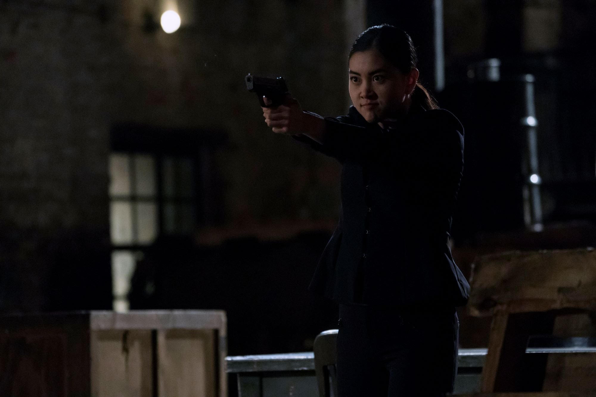 'The Blacklist' star Laura Sohn, in character as Alina Park, wears a dark jacket and pants and points her gun.