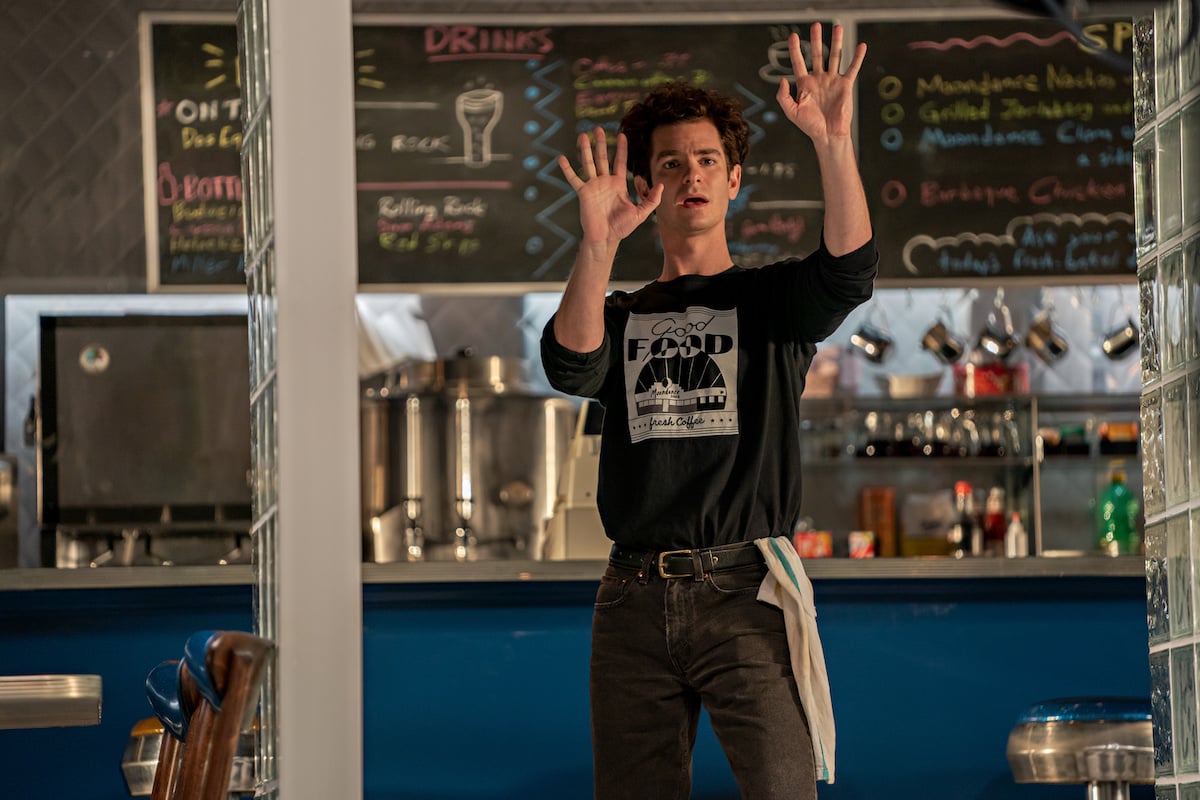 Andrew Garfield as Jonathan Larson in 'Tick, Tick...BOOM!' He stands in a diner in a black long-sleeved shirt and jeans with his arms and hands stretched out in front of him as he imagines something.