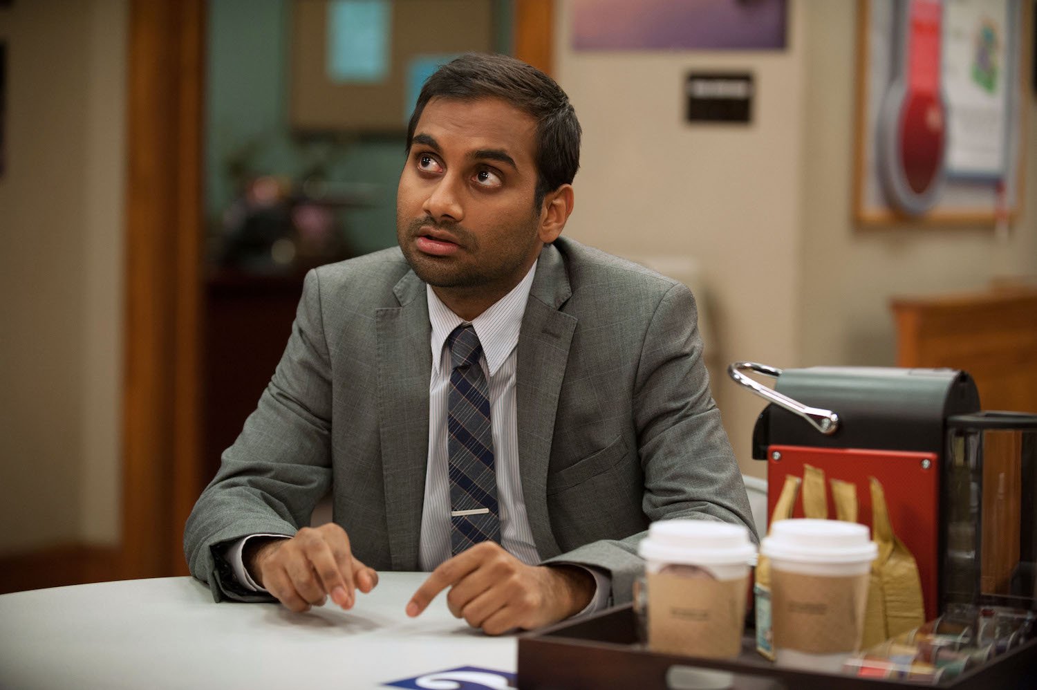 Aziz Ansari sits at a desk in Parks and Recreation